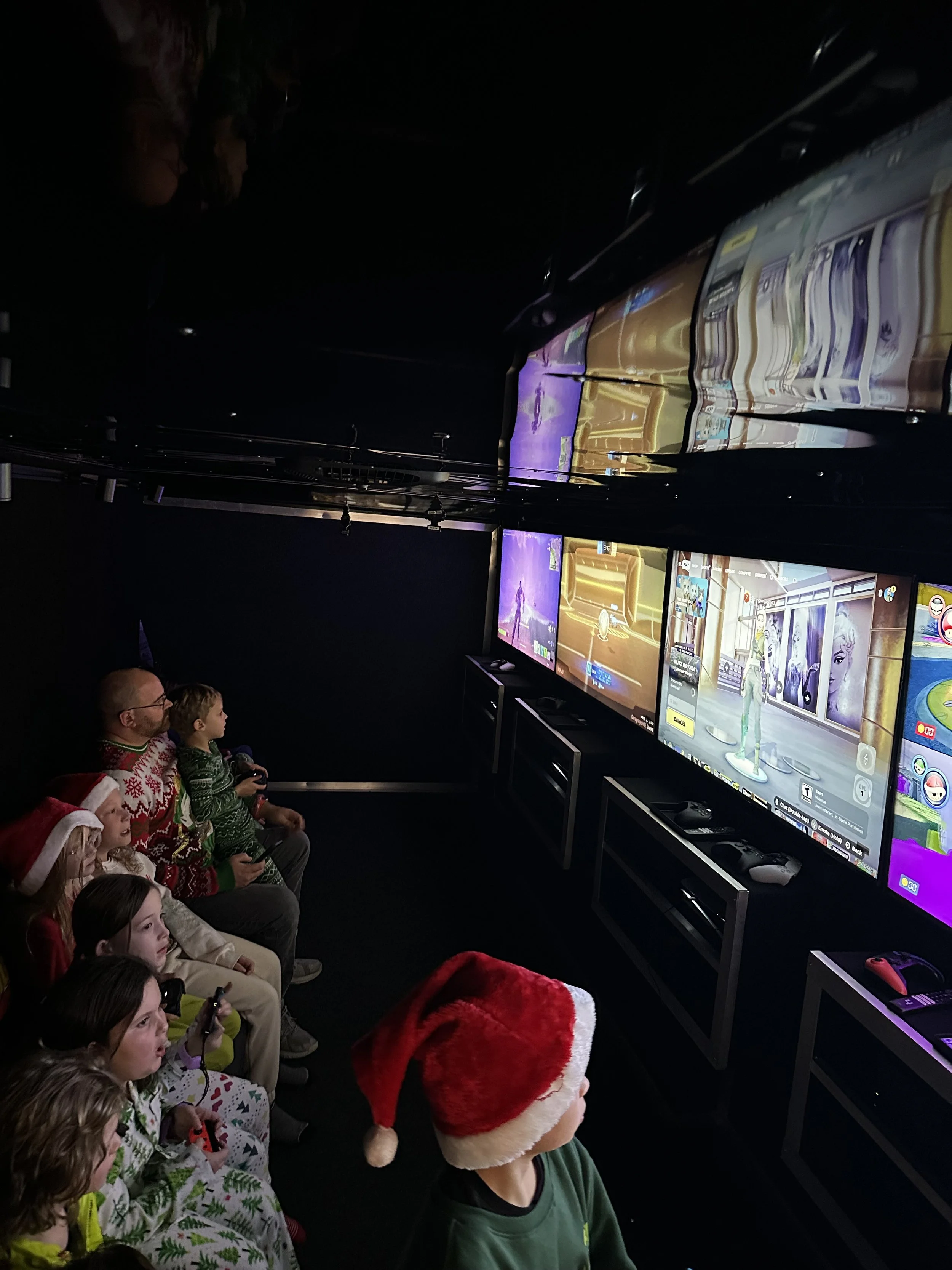 Children and adults wearing Christmas sweaters and Santa hats playing video games on multiple screens in a dark gaming room for a video game Christmas party.