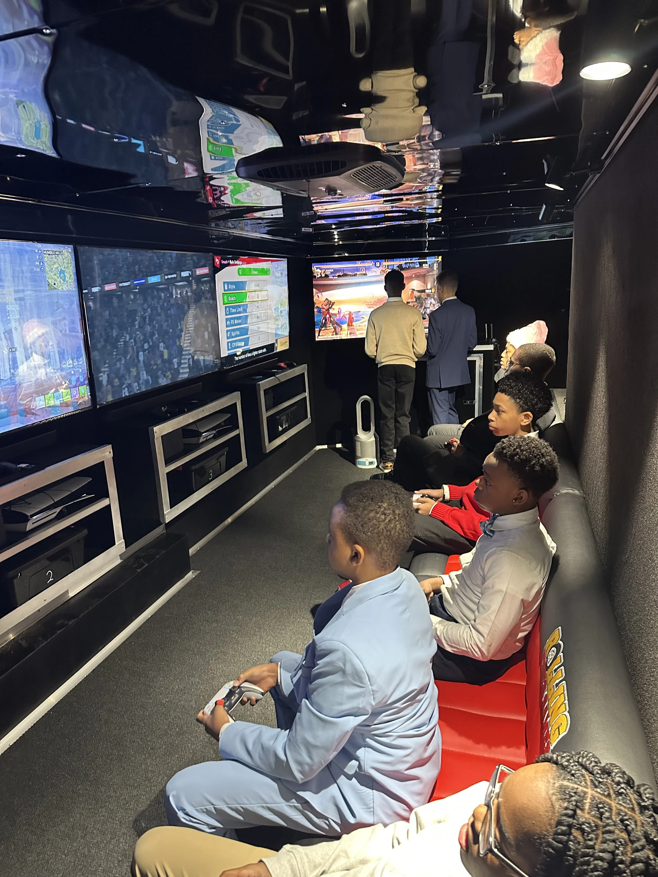 Young boys playing video games in an gaming lounge with large screens, some seated and some standing, with a few people watching.