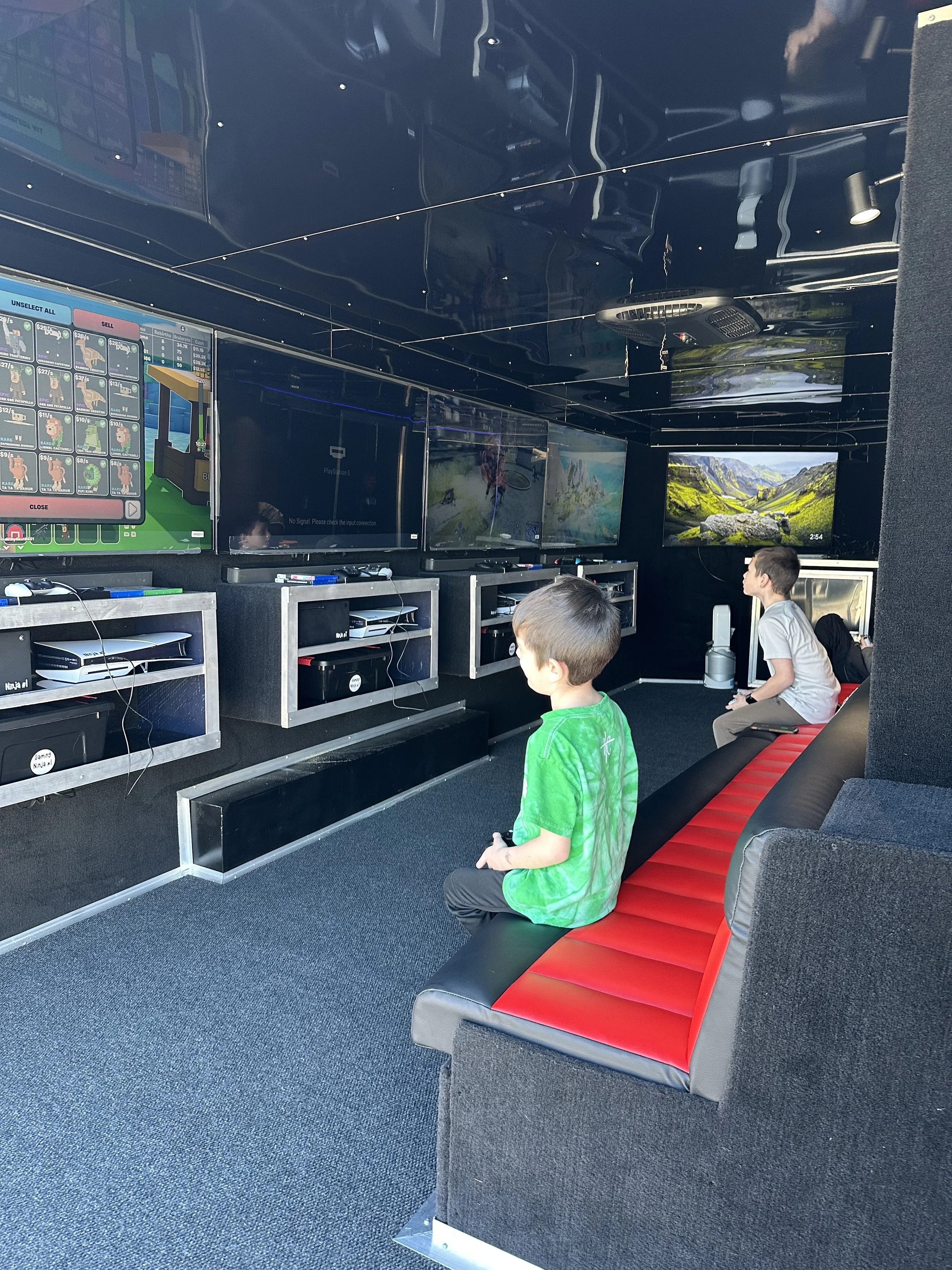 Two young boys sitting on a black and red leather bench in a gaming truck, facing multiple large flat-screen monitors displaying video games and scenic screens, with gaming consoles below the screens for the best birthday idea party ever.