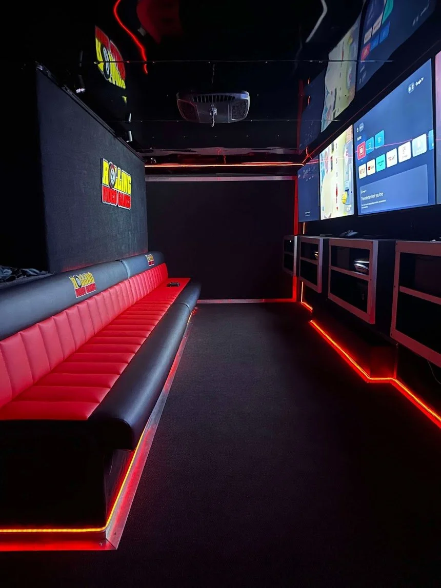 A video gaming trailer for church events and birthdays with a long black and red bench seat along the left wall and multiple large screens on the right wall. The room is illuminated with red LED strip lighting along the floor and ceiling edges.