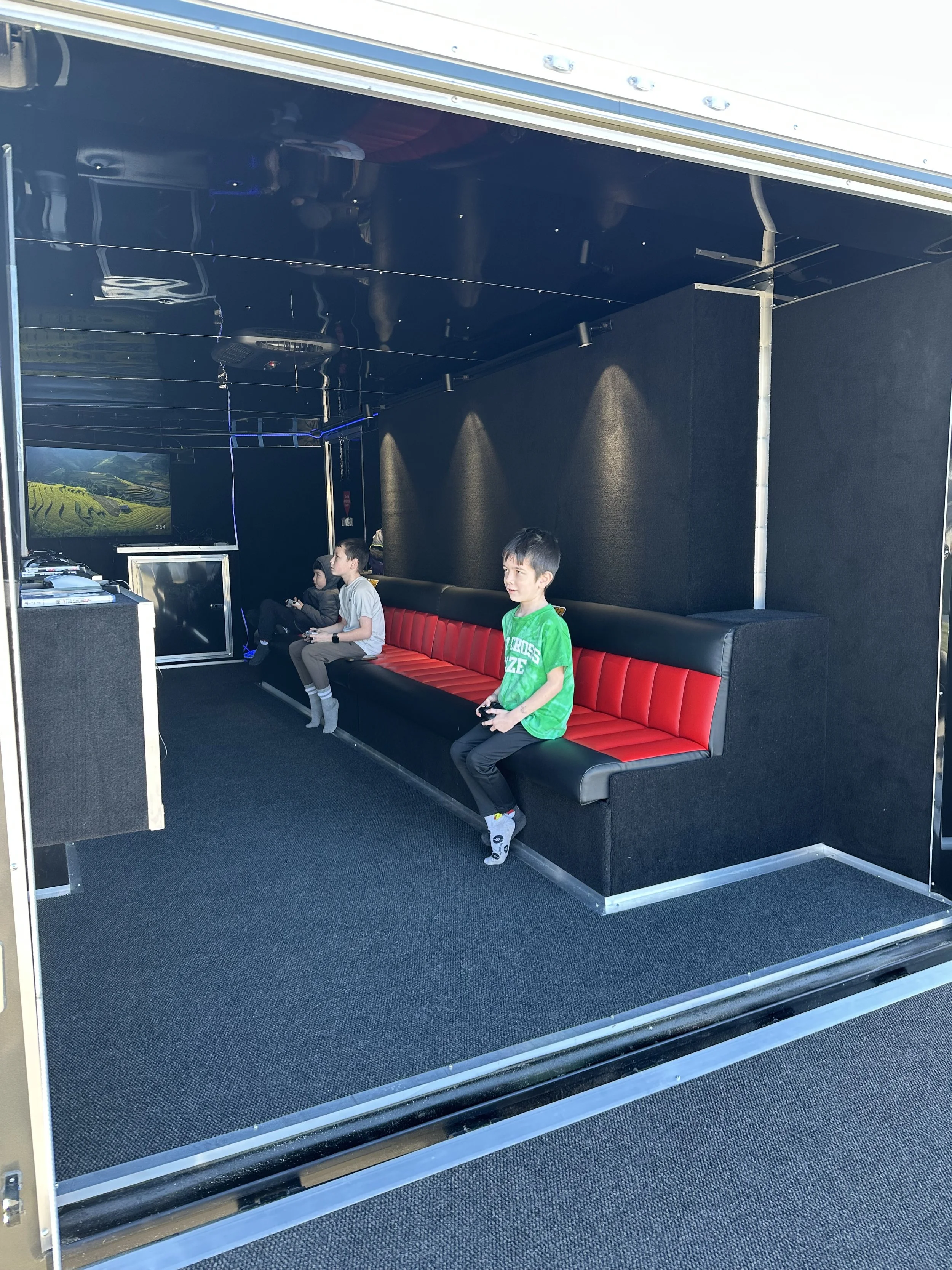 Inside a mobile gaming room with three children playing video games on controllers, sitting on a black and red bench.