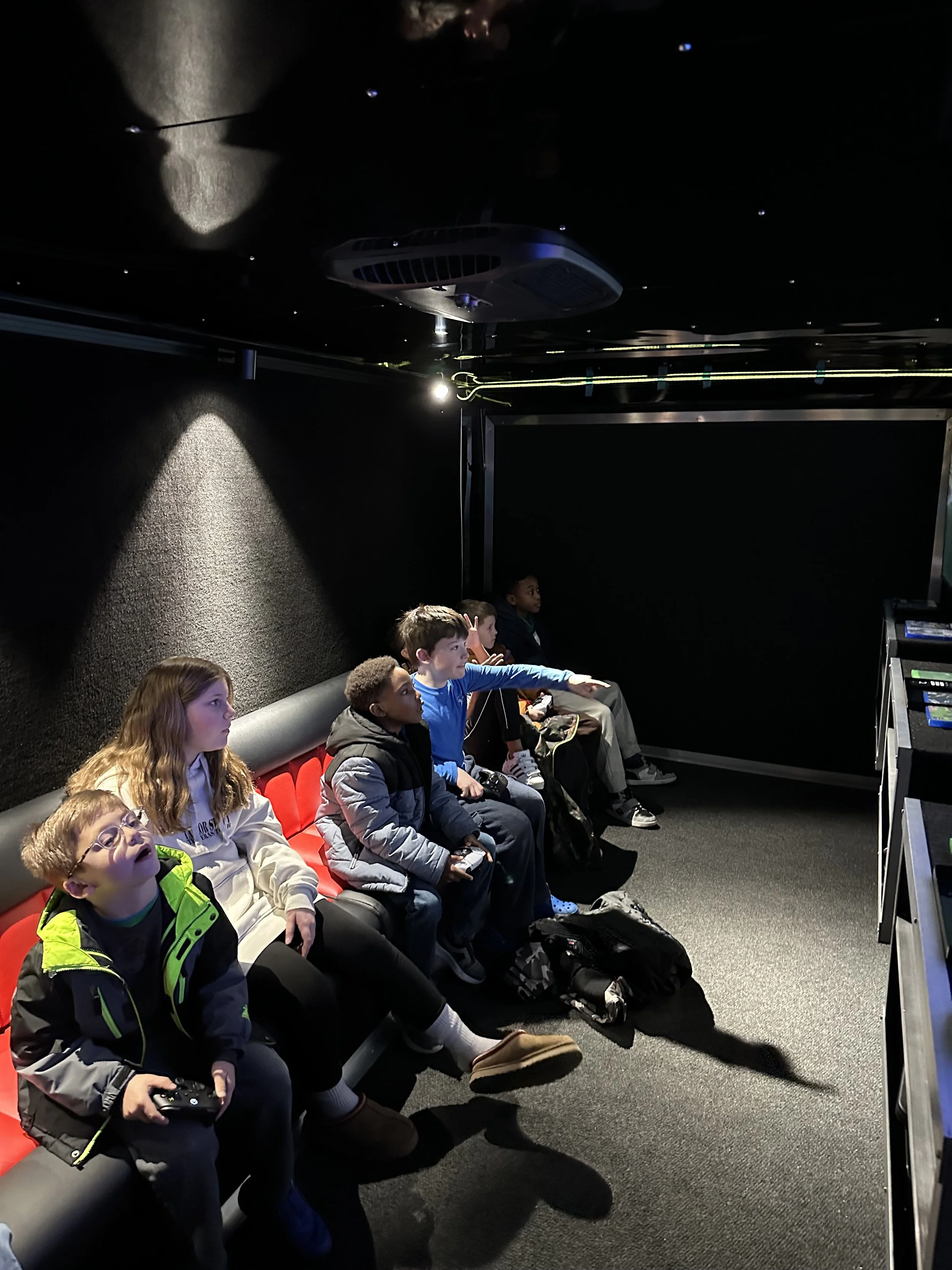Group of children sitting in a mobile gaming truck, some holding game controllers, with backpacks and bags on the floor.