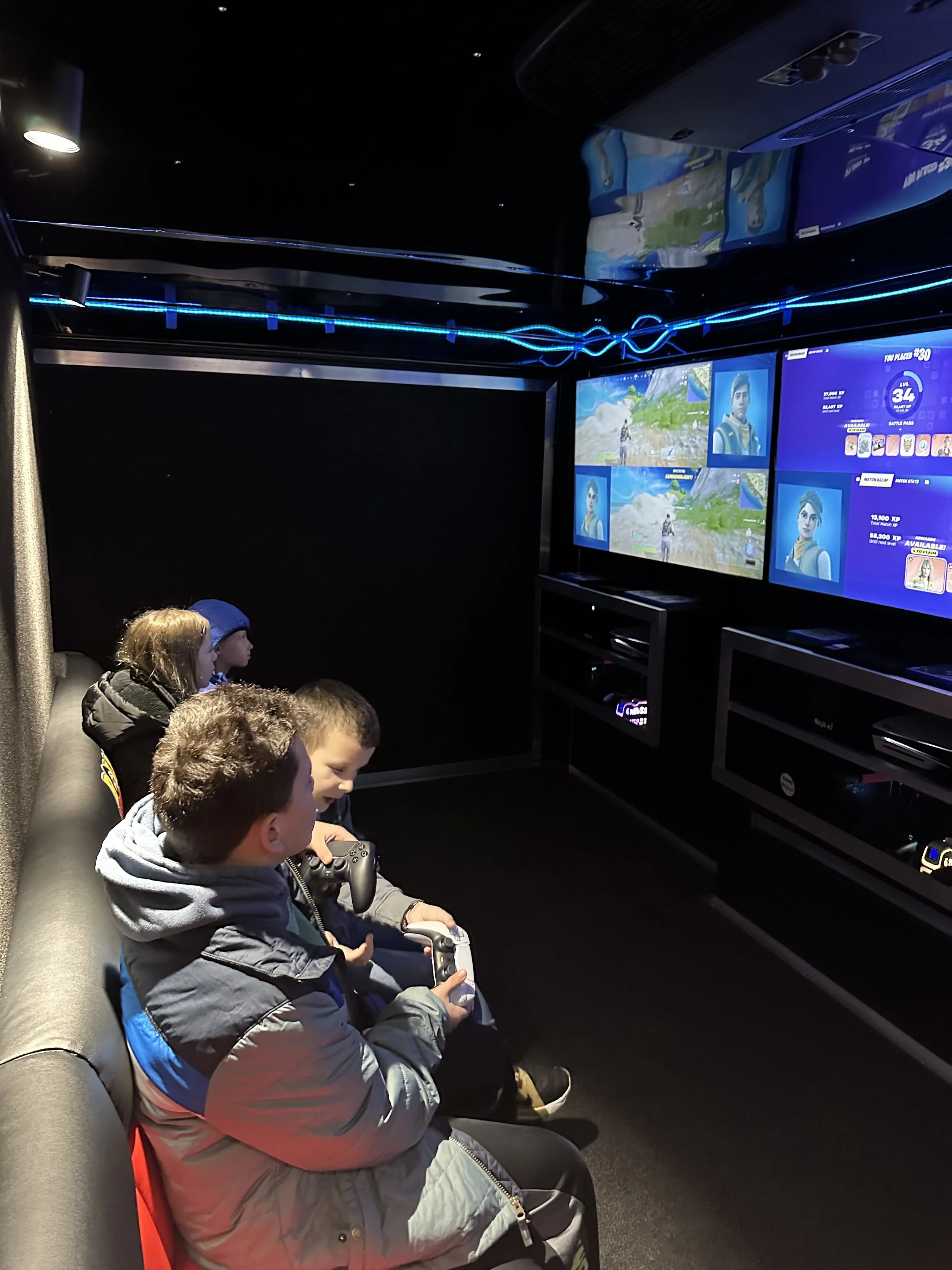 Four children sitting on a couch in front of two large screens, playing a video game with controllers in a mobile gaming trailer adorned with blue LED lights.