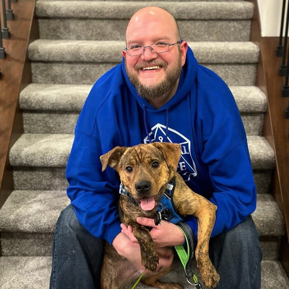 A man with glasses and a beard in a blue hoodie smiling and holding a brindle puppy on a staircase.