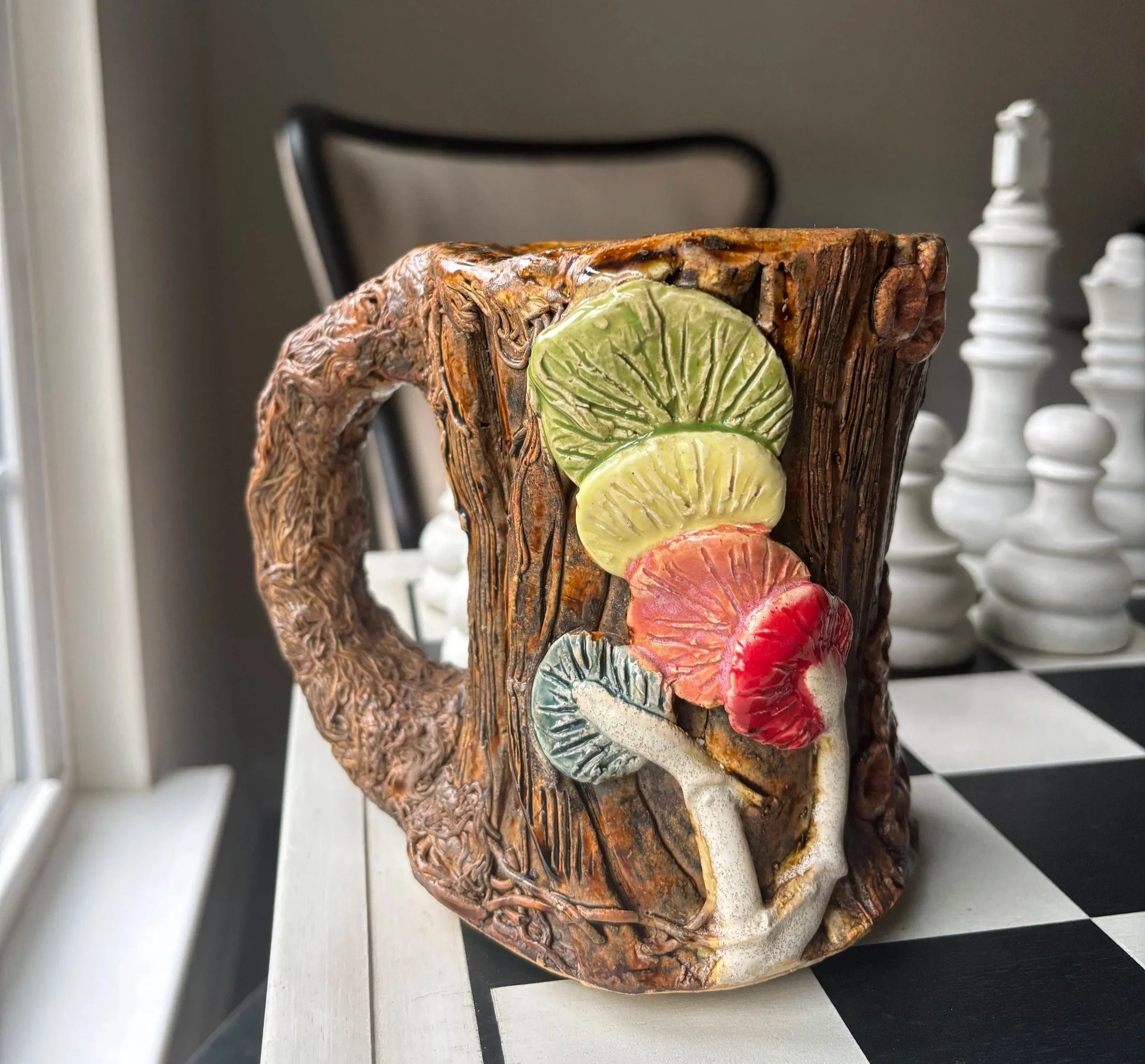 A pottery mug with a textured, bark-like surface featuring colorful mushrooms and a small leaf design. Background includes a chessboard and chess pieces.