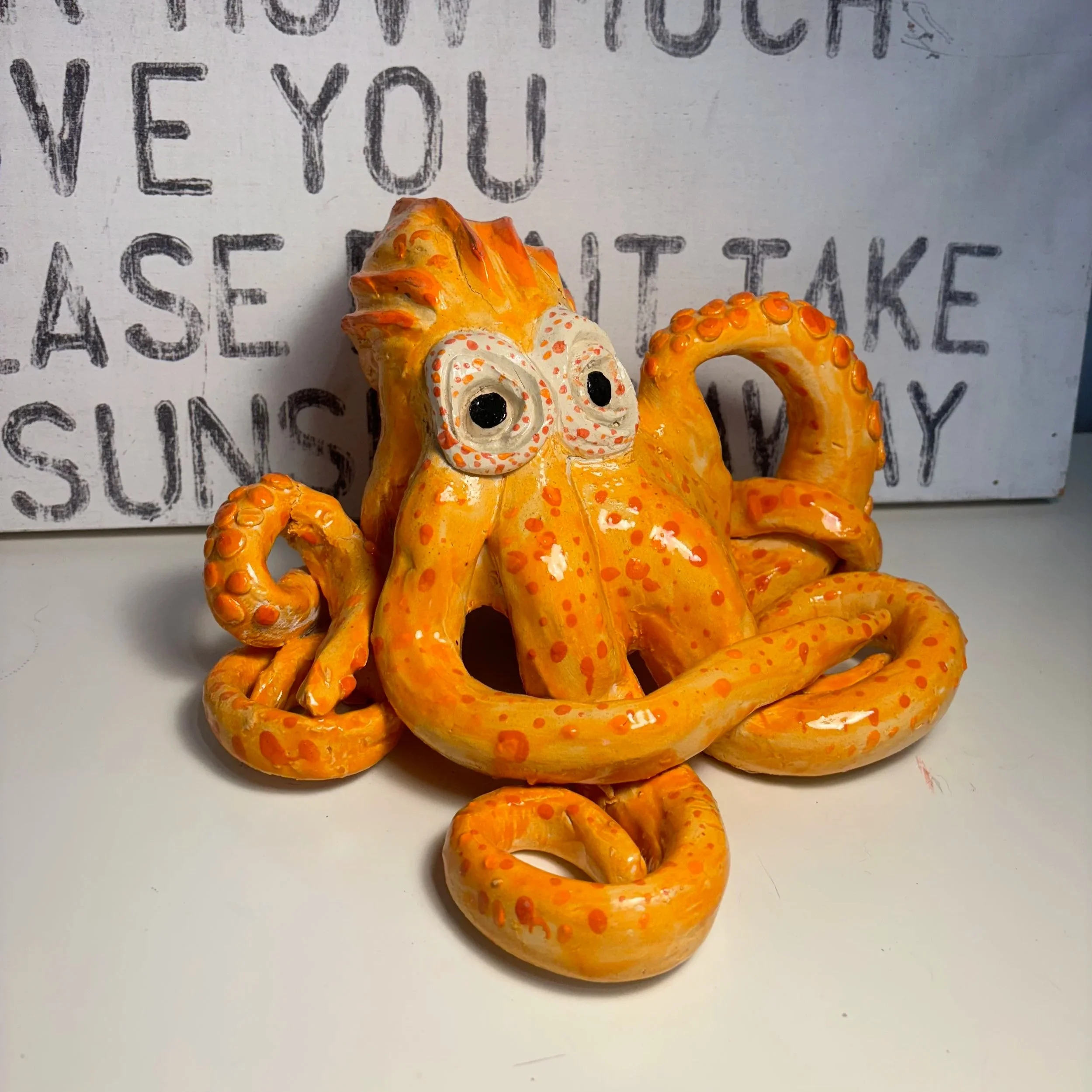 Orange and yellow ceramic octopus sculpture with large eyes, three prominent tentacles, and a textured body, placed on a white surface with a painted sign in the background.