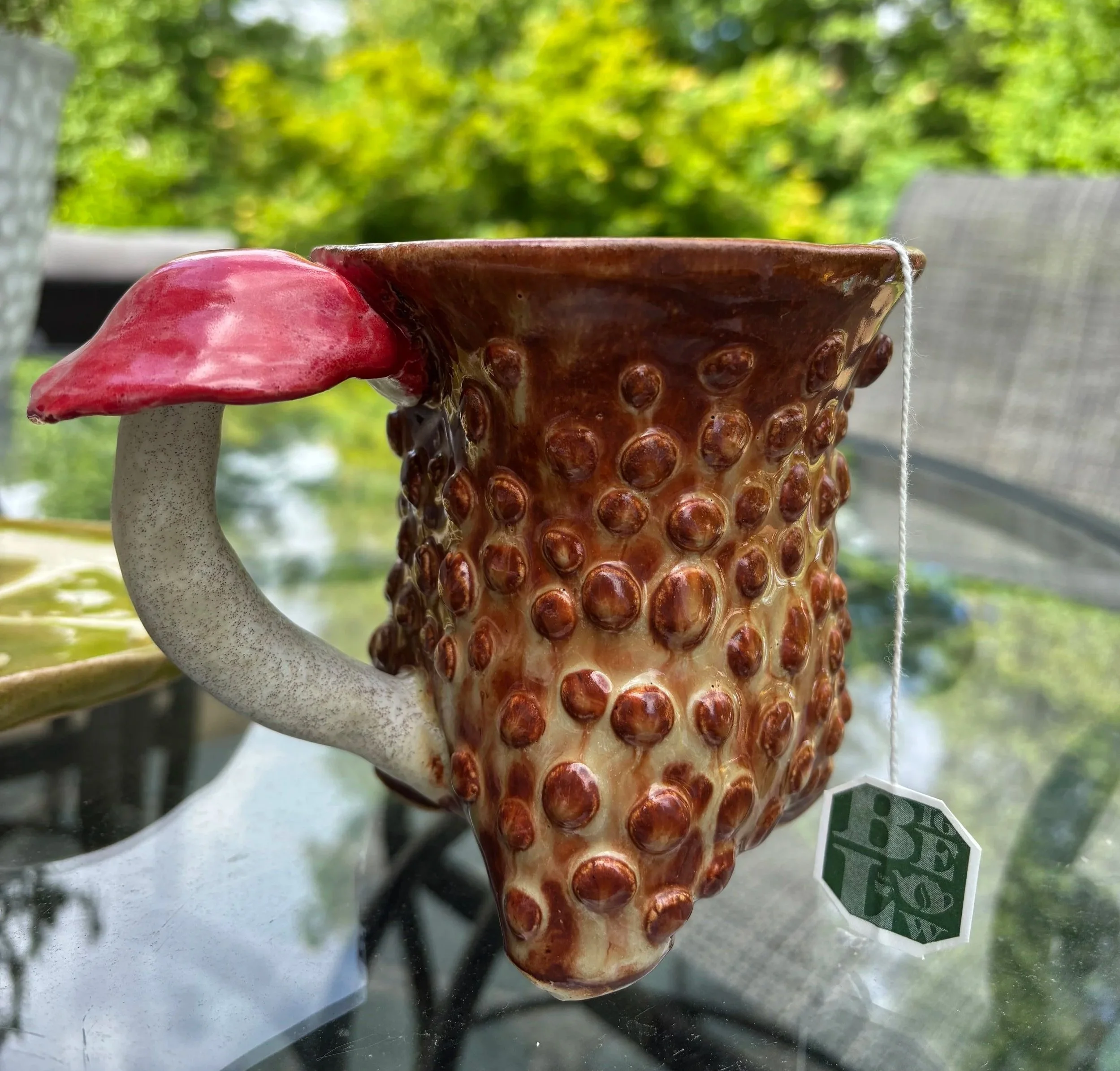 Ceramic mug with textured, bumpy surface resembling a strawberry, featuring a red mushroom-shaped handle, set outdoors with green foliage in the background.
