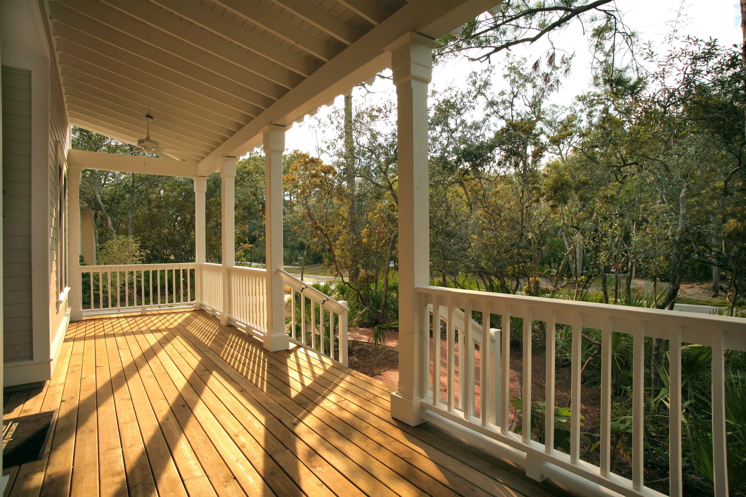Wooden porch with white railings and support columns, overlooking a lush green landscape with trees and a brick pathway.
