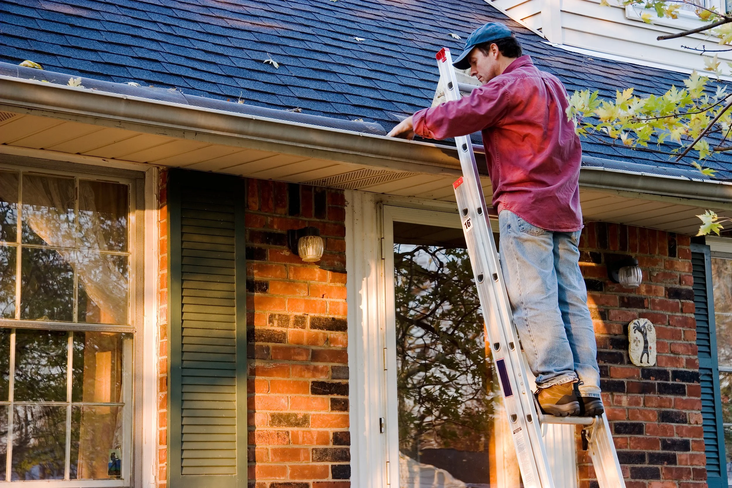 A man standing on a ladder working on the eaves or roof of a brick house, with trees visible in the background.
