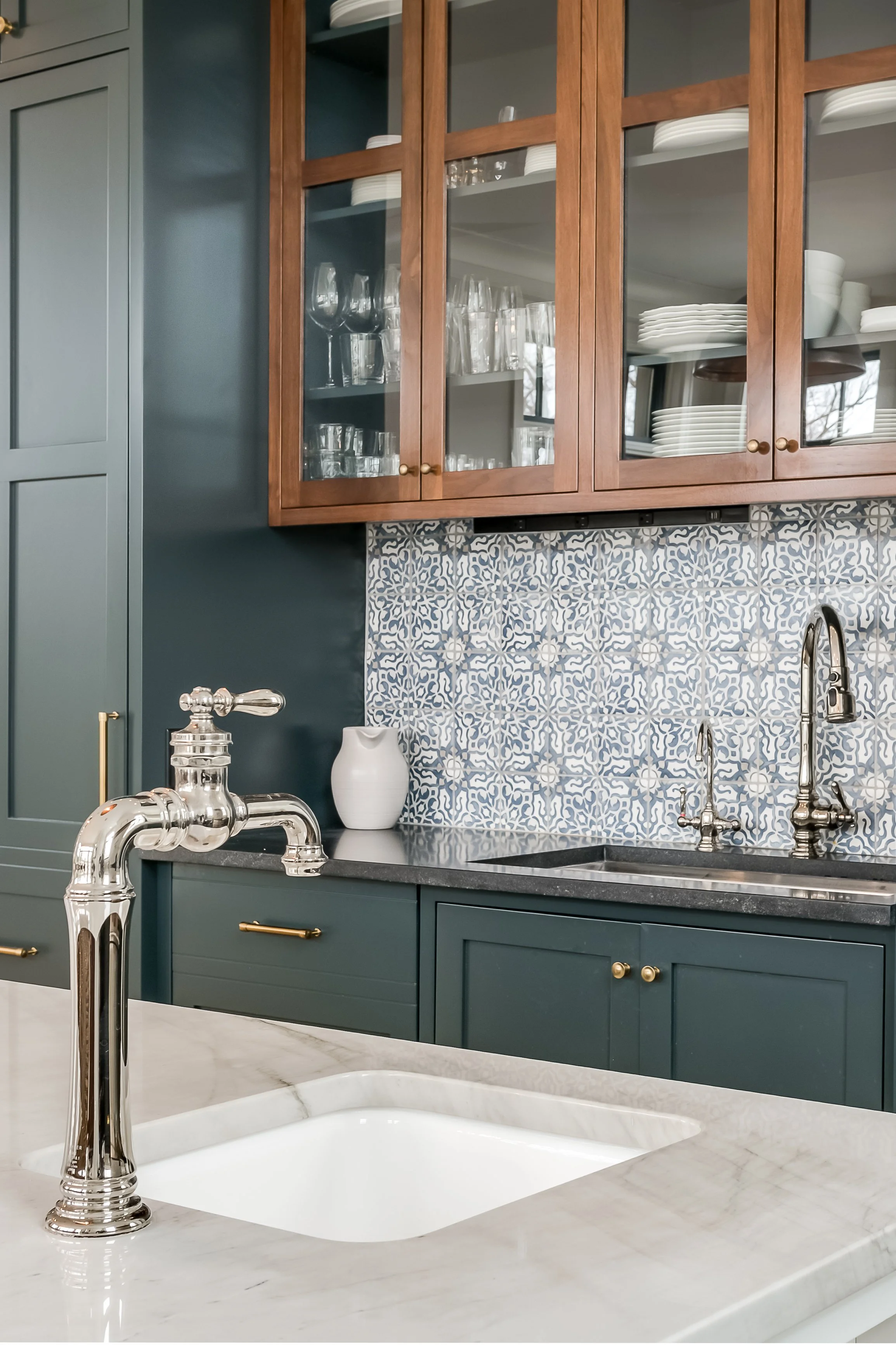 Close-up of a kitchen with a marble countertop, a polished silver faucet, dark green lower cabinets with gold handles, a white vase on purple countertop, patterned blue and white tiles, and glass-front wooden upper cabinets with plates and glassware.