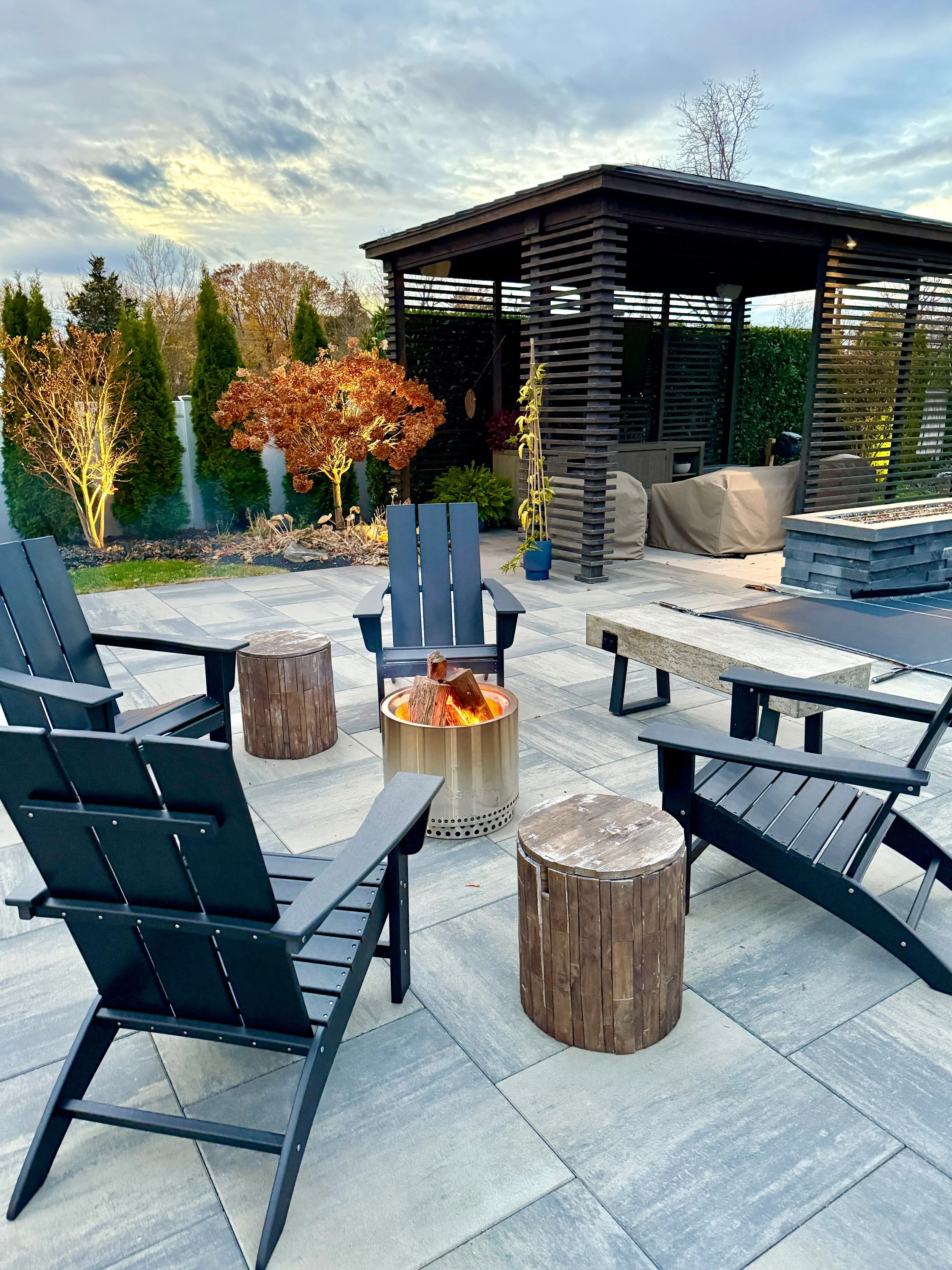 Outdoor patio with four black chairs arranged around a fire pit with burning logs, two wooden stools, and a modern pergola in the background. There are landscaped trees and a shed or small structure with seating and greenery.