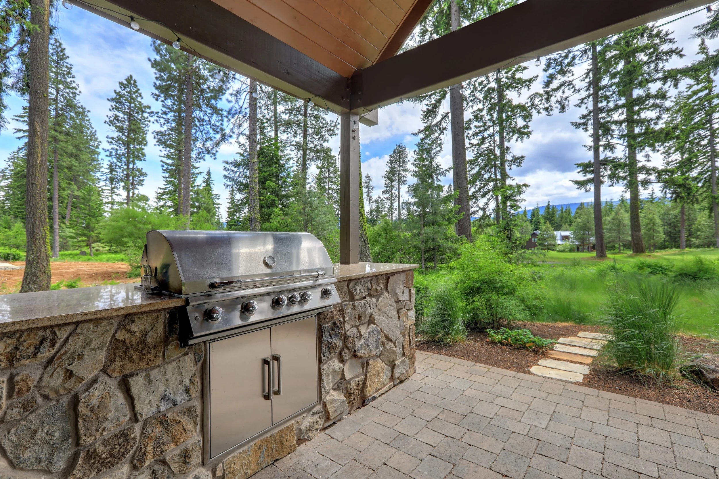 Outdoor kitchen with a stainless steel grill built into a stone counter, surrounded by a lush green forest and a stone pathway.