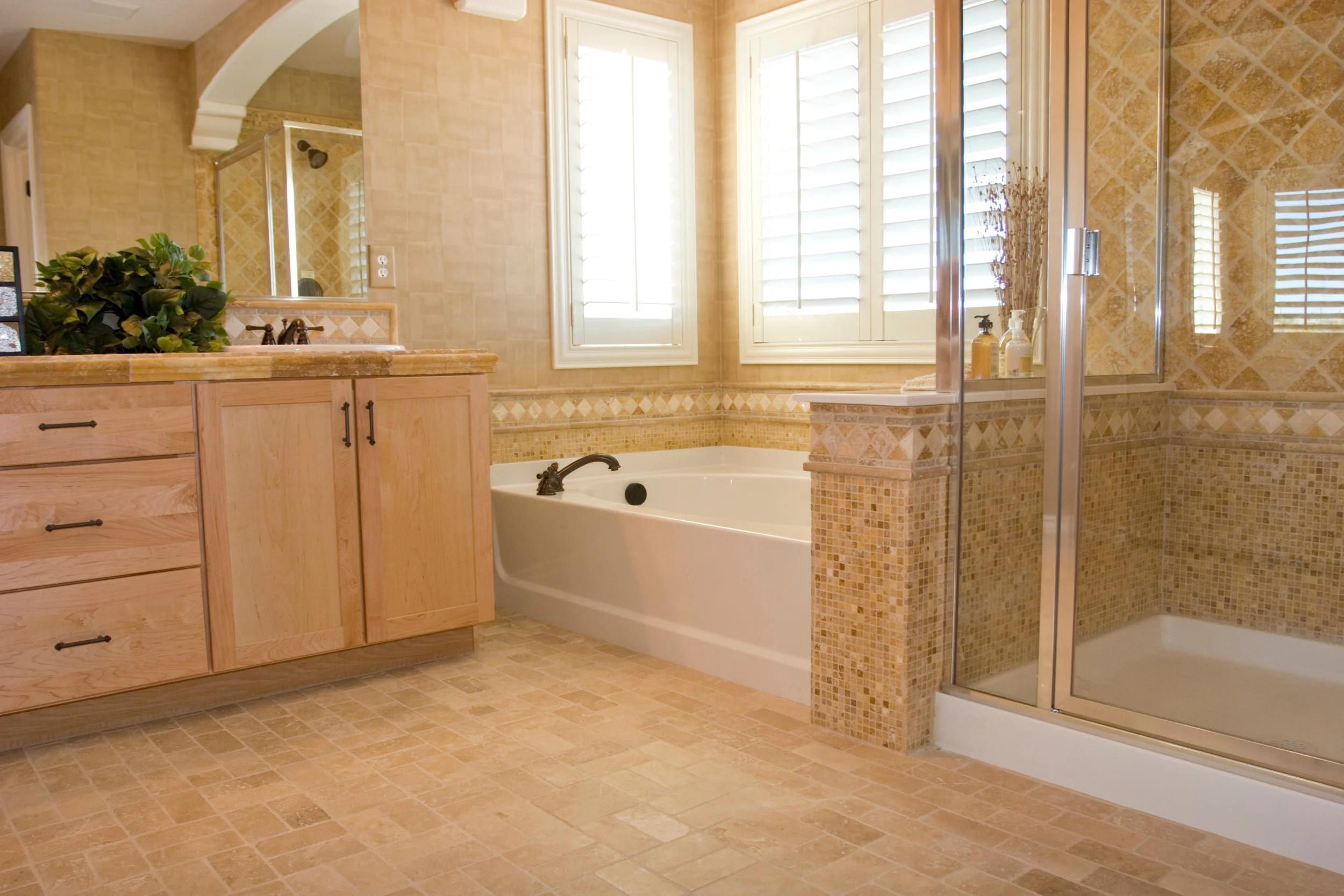 Bathroom with tub, glass shower, wooden vanity, mosaic tile accents, shutters on windows, and beige stone flooring.