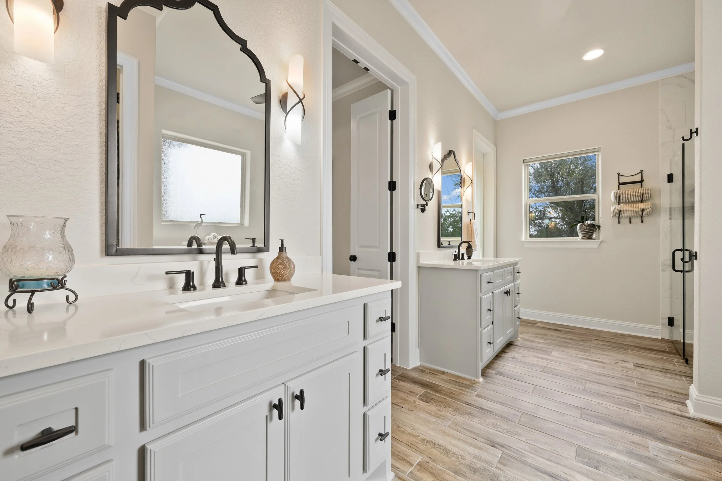 A modern, bright bathroom with two white vanities with black fixtures, large mirrors, and wall-mounted lights. The bathroom has wood-look tile flooring, a window with a view of trees, and a small towel rack with rolled towels. There is a glass shower door in the corner.