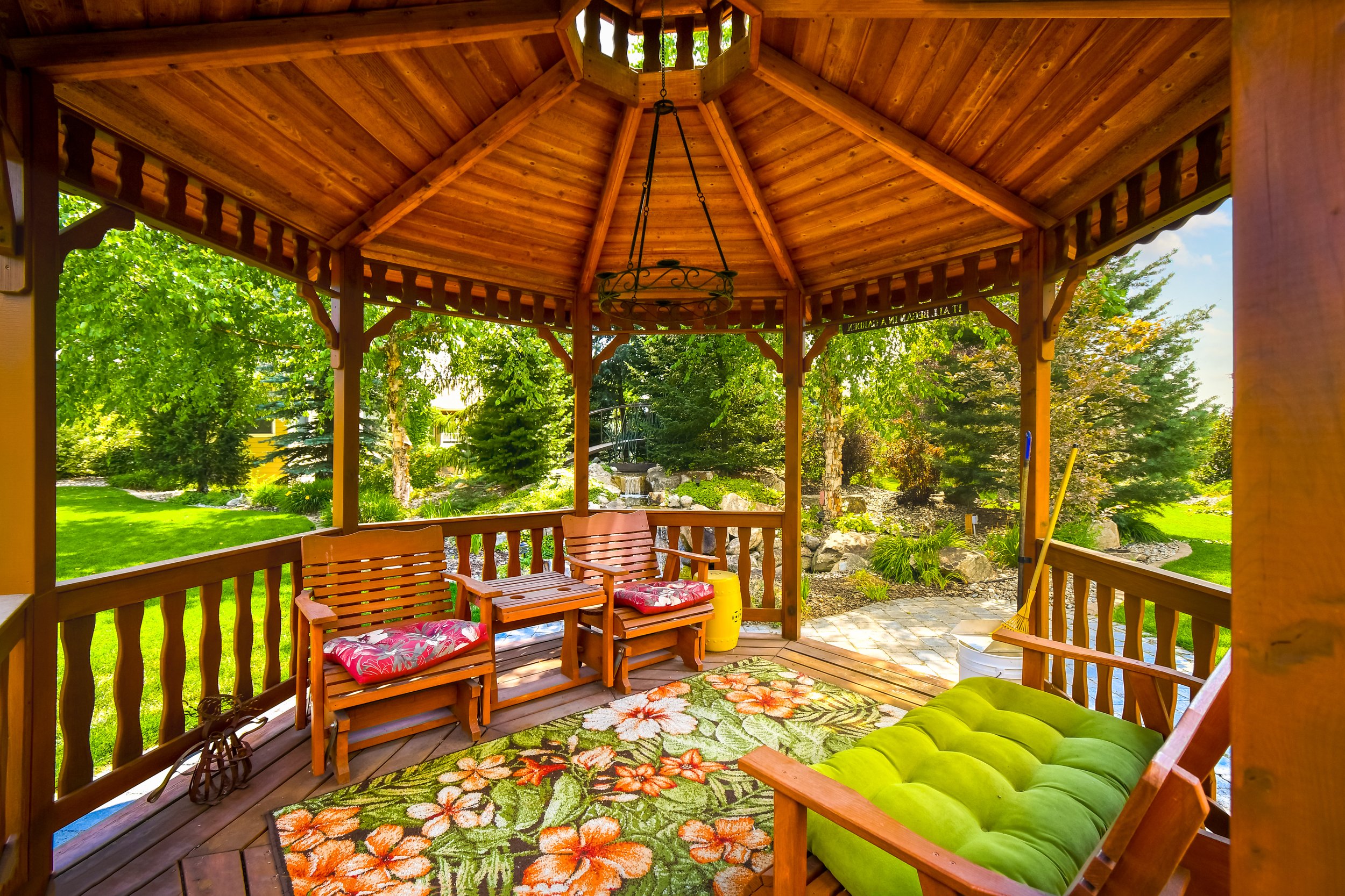 Covered wooden porch with green and floral cushions on benches, a floral rug, surrounded by greenery, trees, and a landscaped garden with rocks and a small waterfall in the background.