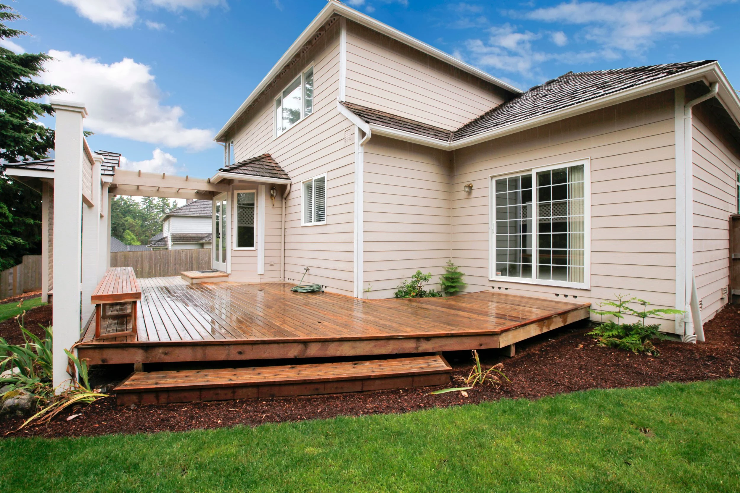 Backyard with a wooden deck attached to a two-story house, sliding glass door, and large window, surrounded by a lawn, plants, and a fence under a partly cloudy sky.