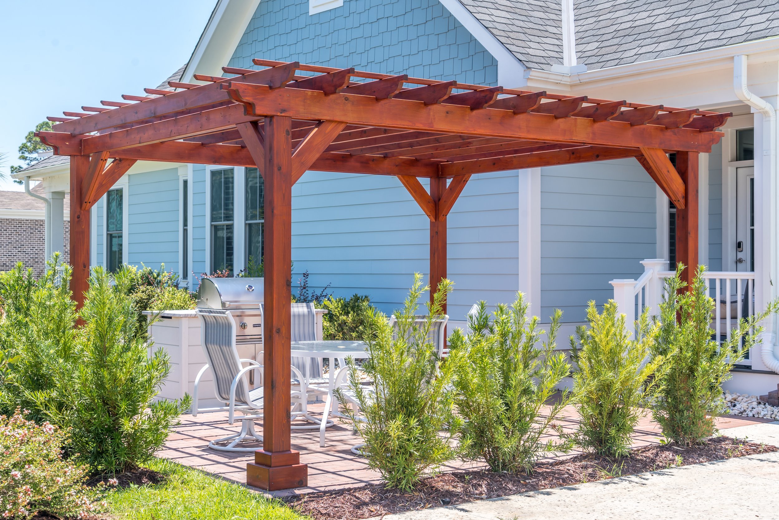 A backyard patio with a wooden pergola, outdoor table, chairs, a barbecue grill, and surrounding greenery, attached to a light blue house with white trim.