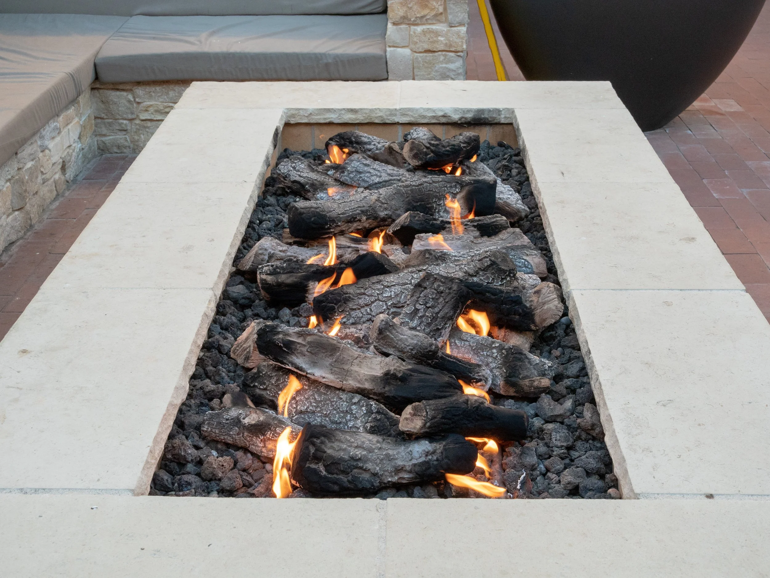 Outdoor fireplace with black lava rocks and flames, surrounded by a beige stone border.