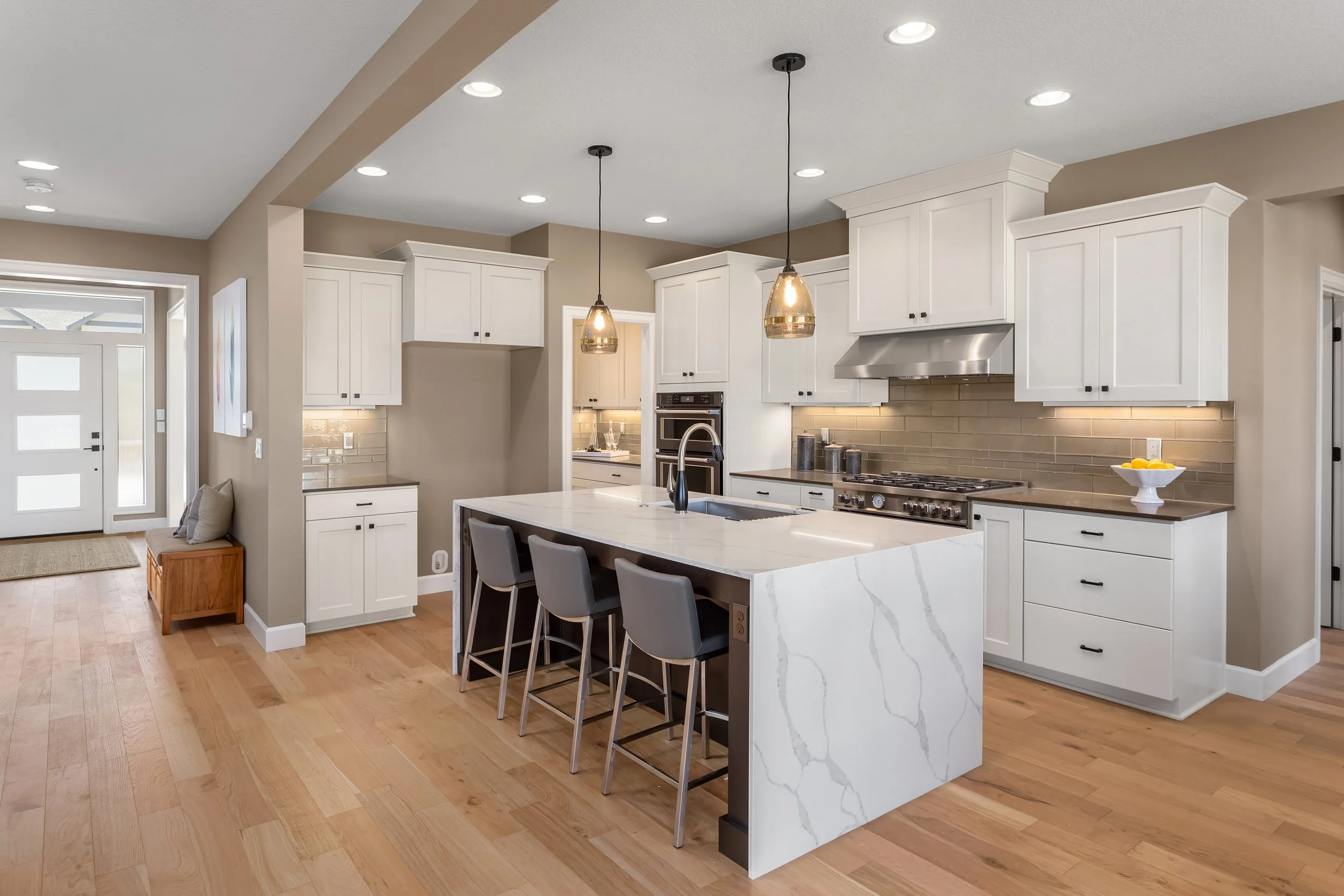 Modern kitchen with white cabinets, a marble island with a built-in sink, pendant lighting, and stainless steel appliances. Hardwood floors and a door leading outside are visible.