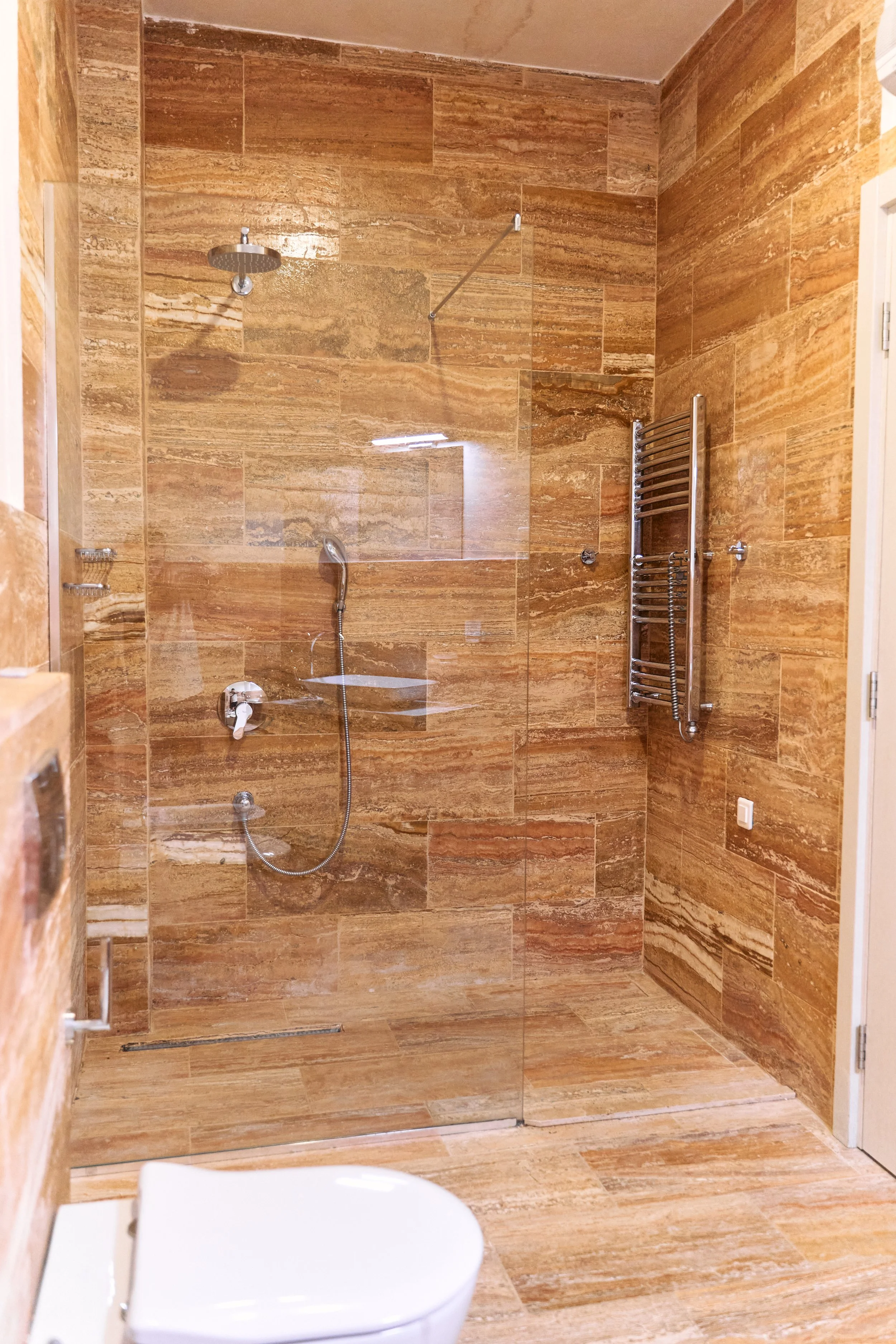 A modern bathroom with a glass-enclosed walk-in shower featuring brown marble tiles, a rain showerhead, a handheld shower, a heated towel rack, and a toilet at the bottom.