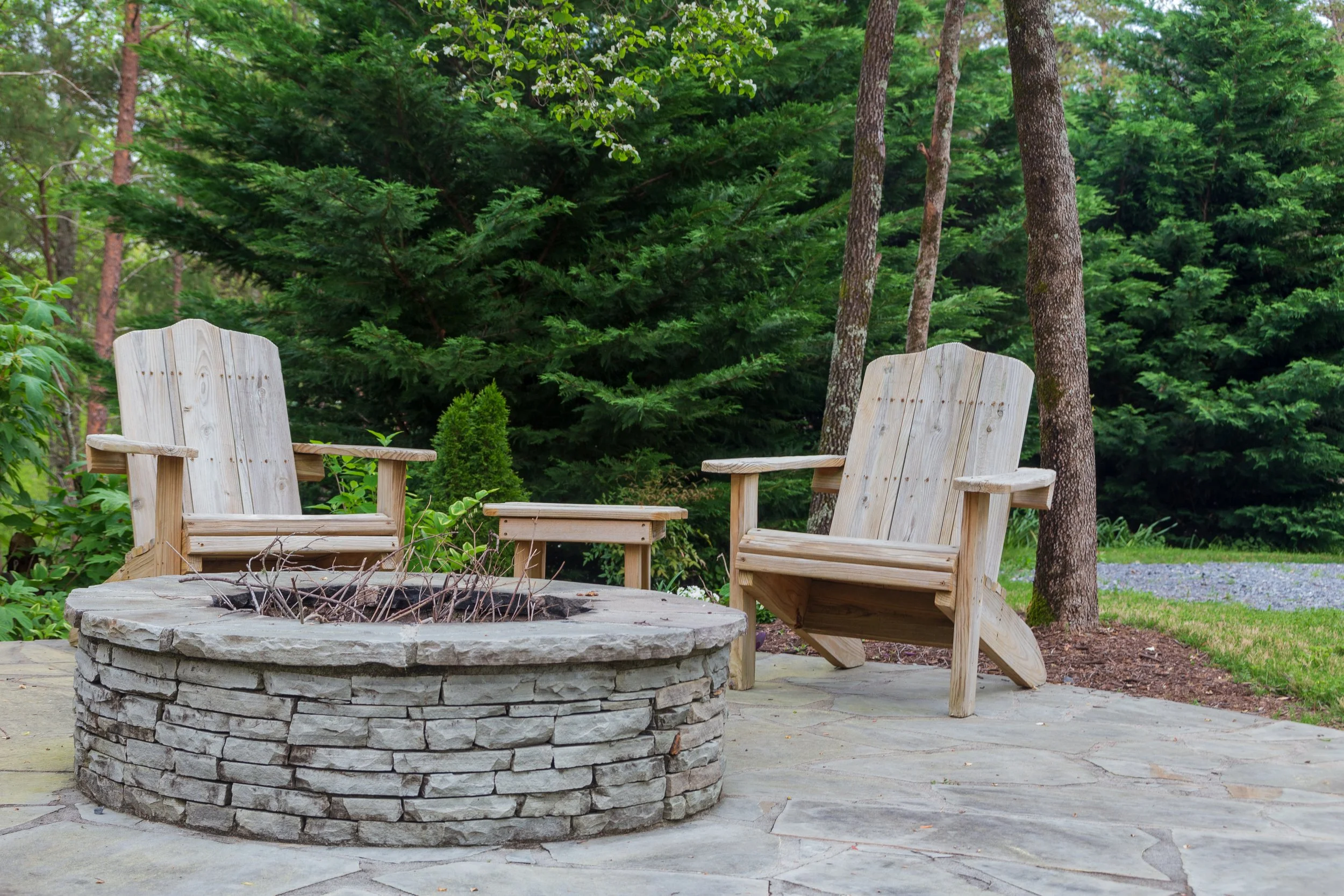 Two wooden Adirondack chairs and a small side table arranged around a stone fire pit on a patio with trees and green foliage in the background.