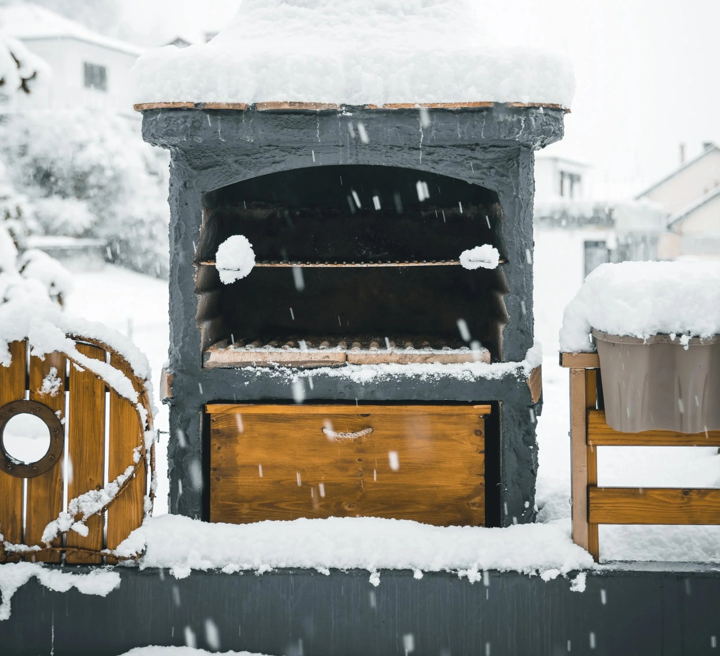 A snow-covered outdoor pizza oven with a wooden cabinet below, flanked by wooden planters also covered in snow, in a snowy backyard.