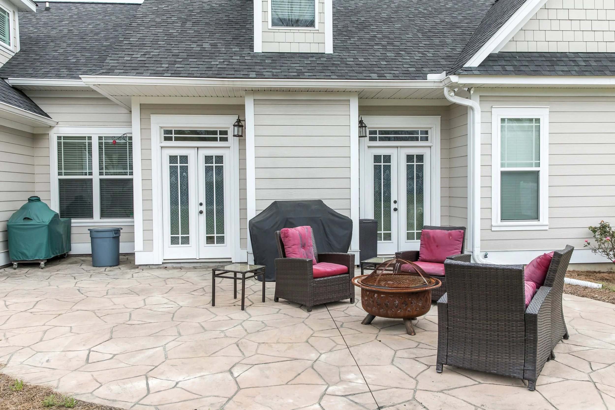 Backyard patio with wicker furniture, pink cushions, a fire pit, grill covered with a black cover, and a white house with multiple windows and glass doors.