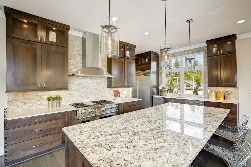 Modern kitchen with dark wood cabinets, stainless steel appliances, a granite island, pendant lights, and a view of a backyard through windows.