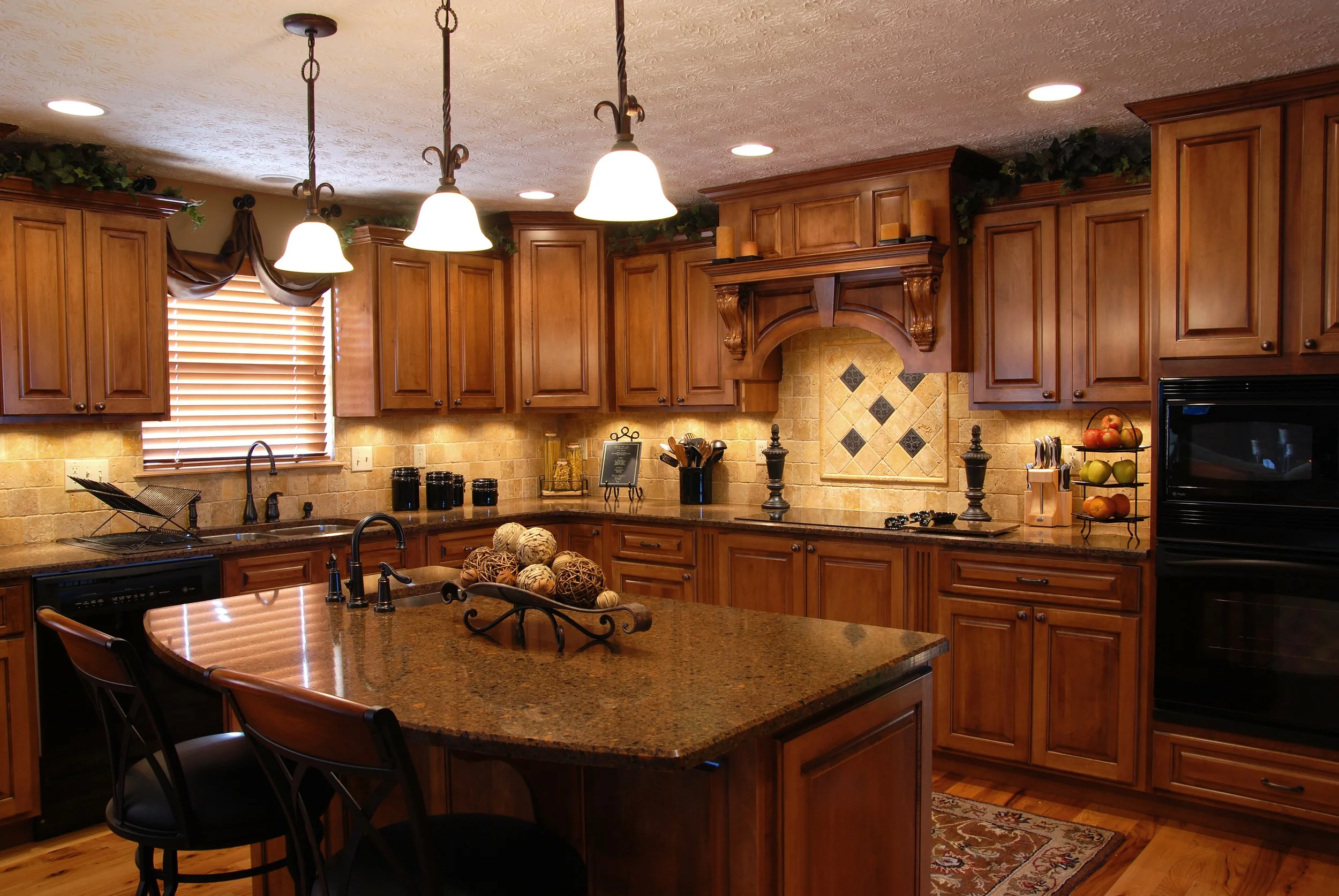 A spacious kitchen with wooden cabinets, a large granite island, black appliances, and warm lighting. Decor includes fruit bowls, a fruit display, and decorative items.