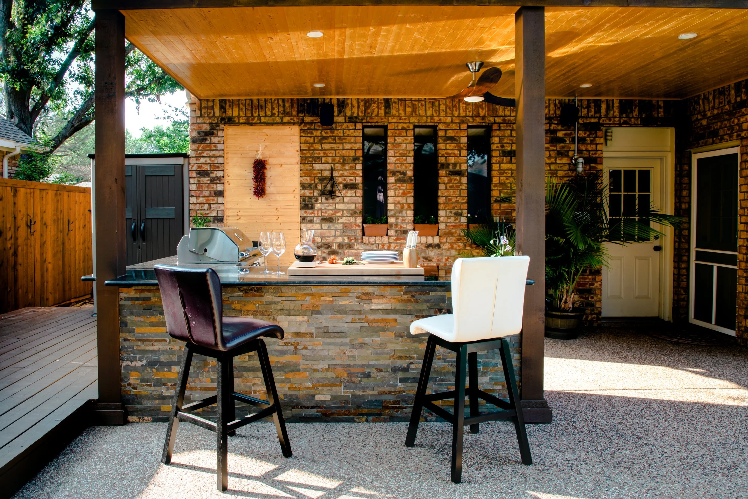 Outdoor patio with a brick wall, wooden ceiling, bar with wine glasses, plates, a pitcher, and a grill, surrounded by wooden decking and greenery.
