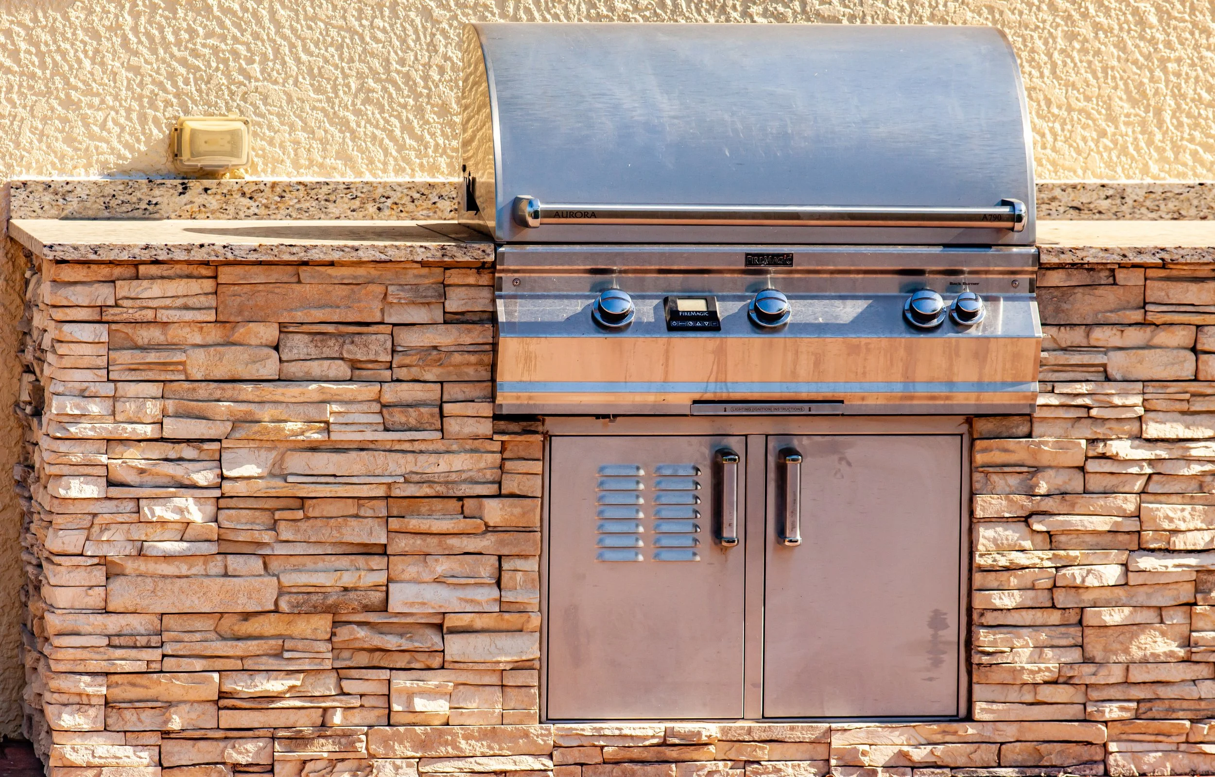 Built-in outdoor grill with stainless steel lid set into a stone and granite countertop against a textured wall.