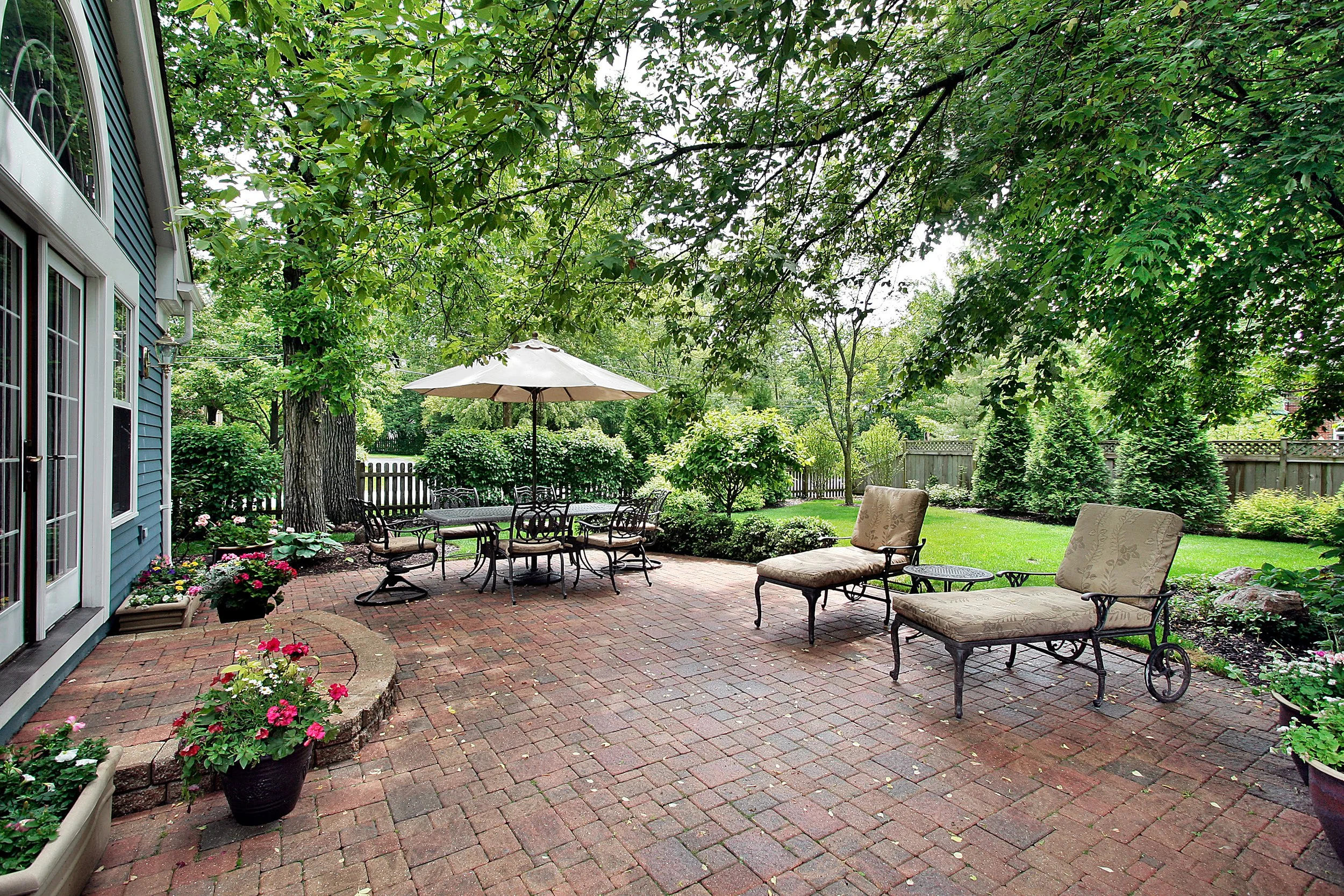 A backyard patio with brick flooring, surrounded by green trees and shrubs. There is outdoor furniture including a black metal dining table with chairs and a large white umbrella, as well as cushioned lounge chairs and small side tables. Potted plants and flowers decorate the area adjacent to the house, which has blue siding and white-framed windows. A wooden fence encloses the yard.