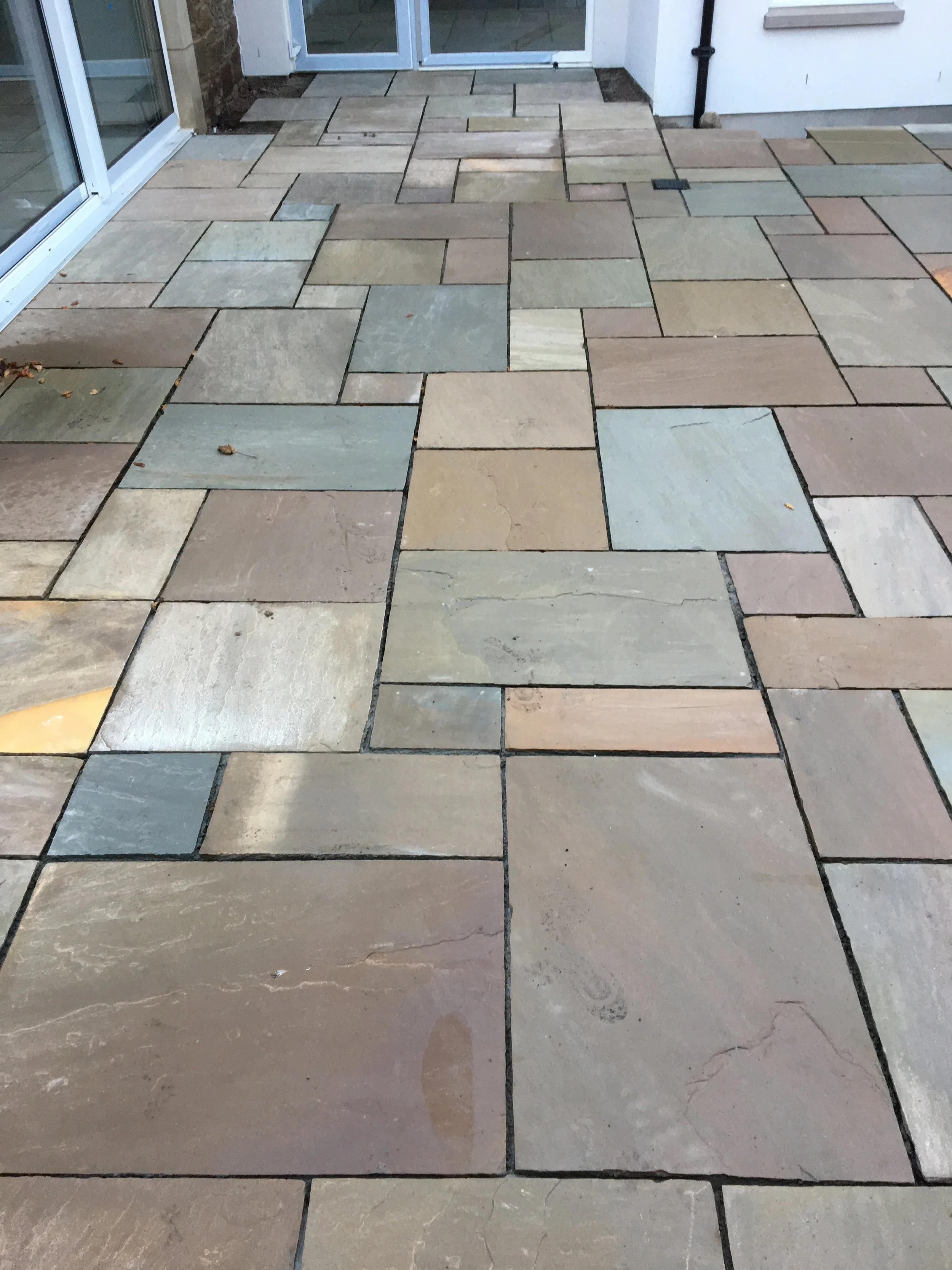 Colorful stone patio with rectangular and square tiles in various shades of beige, gray, and brown, adjacent to a house with white walls and large glass doors.