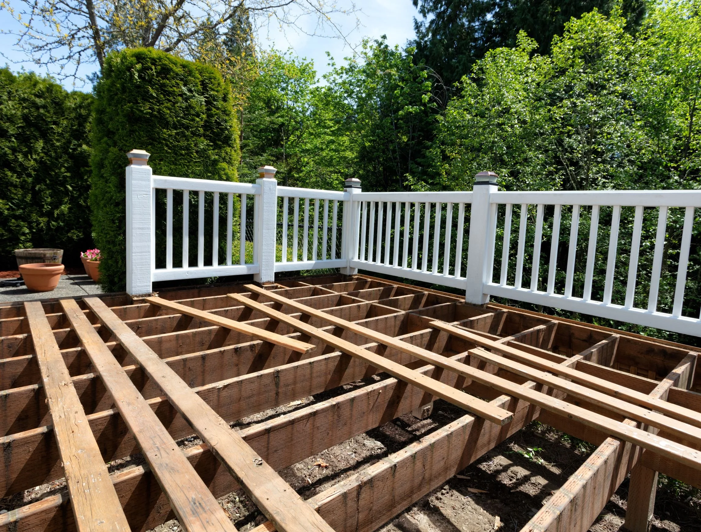 A backyard deck under construction with wooden framing, surrounded by green trees and a white railing fence.