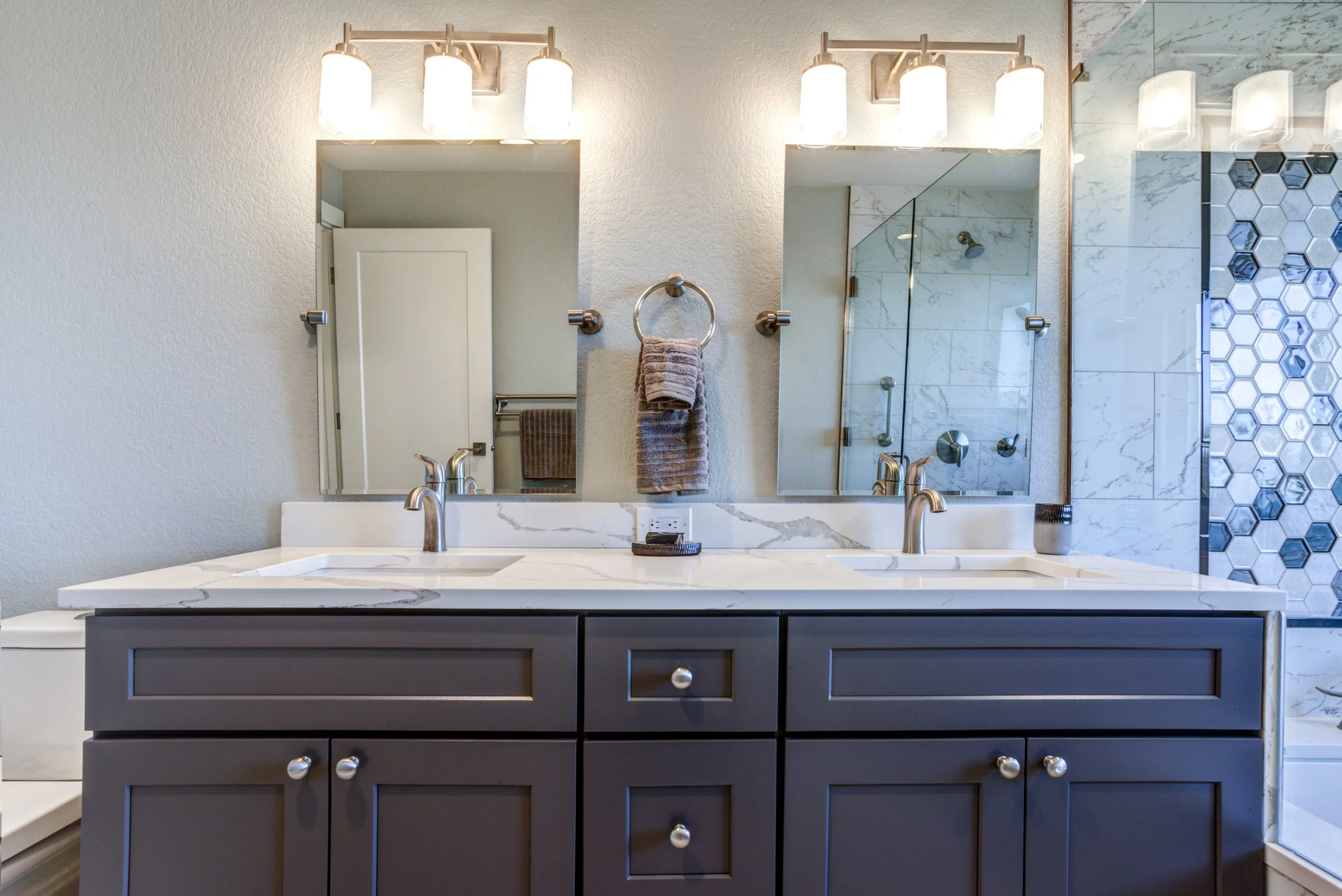 Modern bathroom with a double sink vanity, large mirrors, and contemporary fixtures, including overhead lights, towels on rings, and a walk-in shower with marble wall tiles and hexagonal patterned tile.