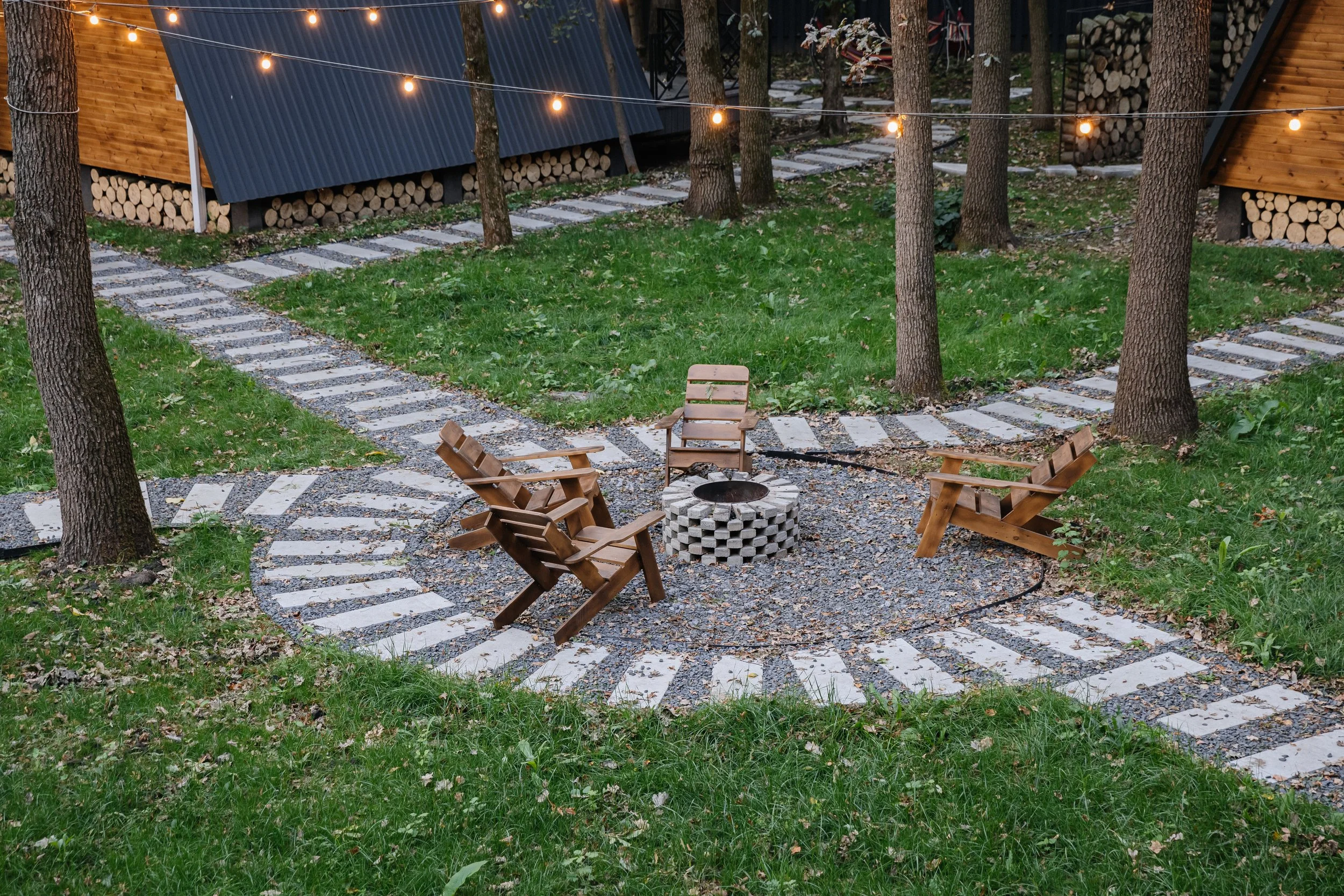 A cozy outdoor fire pit area with four wooden chairs arranged around a circular fire pit made of concrete blocks, surrounded by a gravel and brick paver pathway with a grassy lawn, trees, and string lights overhead at dusk.