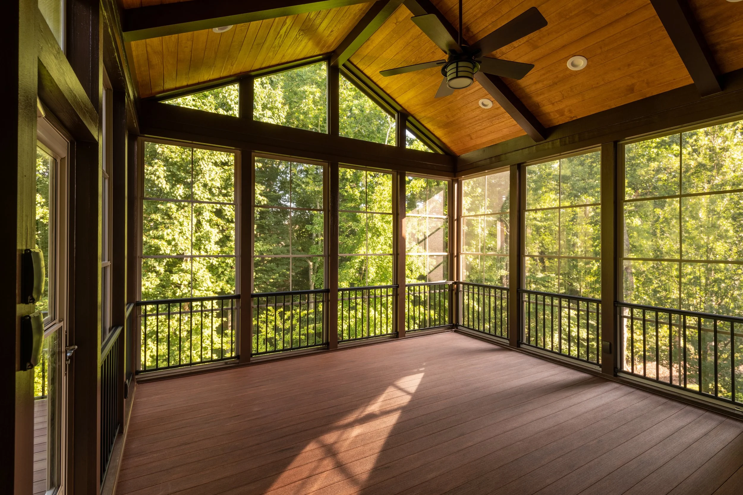 Enclosed porch with large windows, wooden ceiling, ceiling fan, and wooden flooring, overlooking green trees.