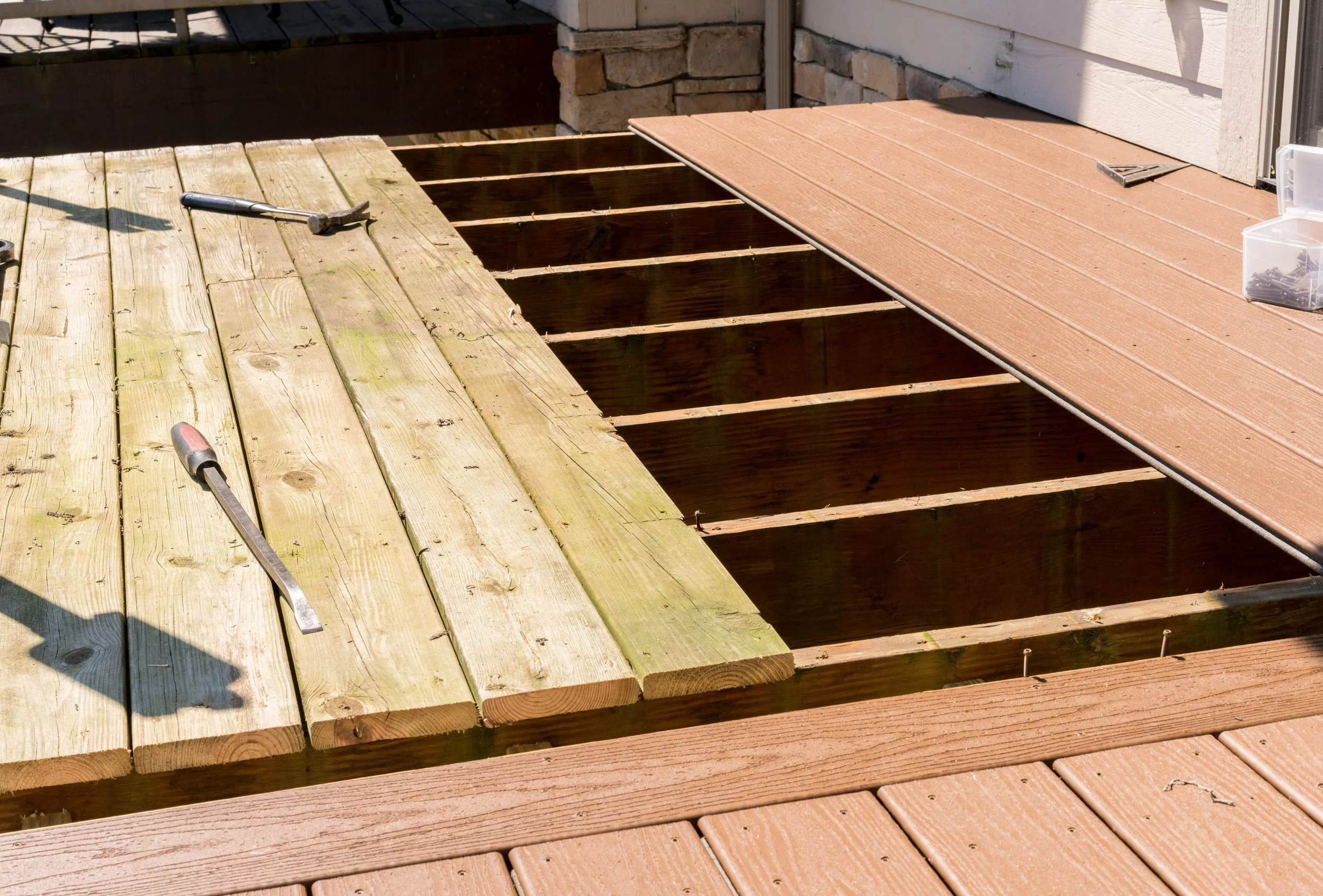 Part of a wooden deck under construction with tools, including a pry bar and hammer, on unfinished and finished sections of the deck.