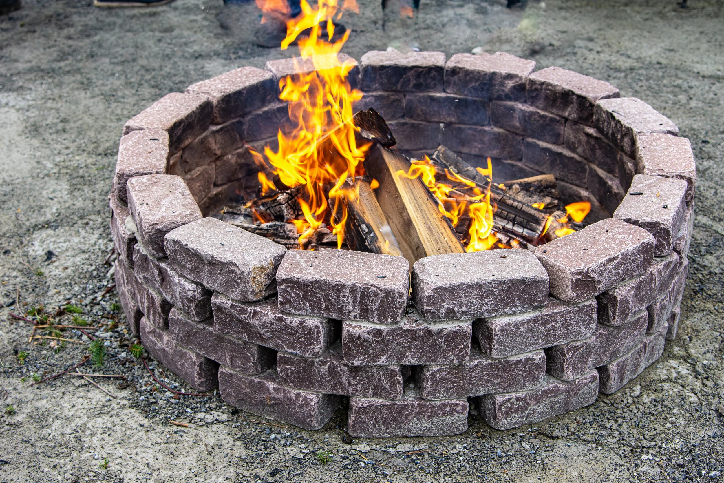 A round fire pit made of stacked bricks with a fire burning inside, sitting on dirt ground.