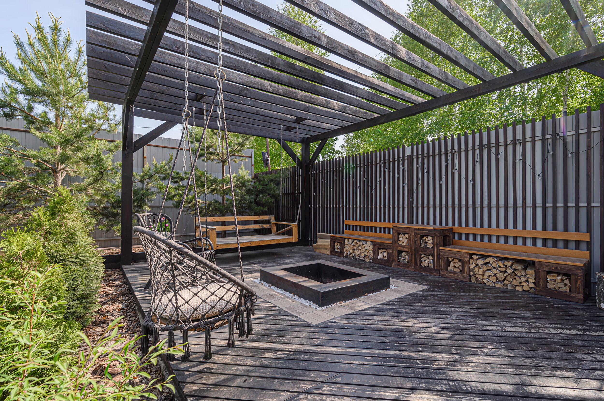 Outdoor patio with wooden decking, a fire pit in the center, a swing and a bench with wood storage underneath, and string lights along the fence. There are trees and greenery around.