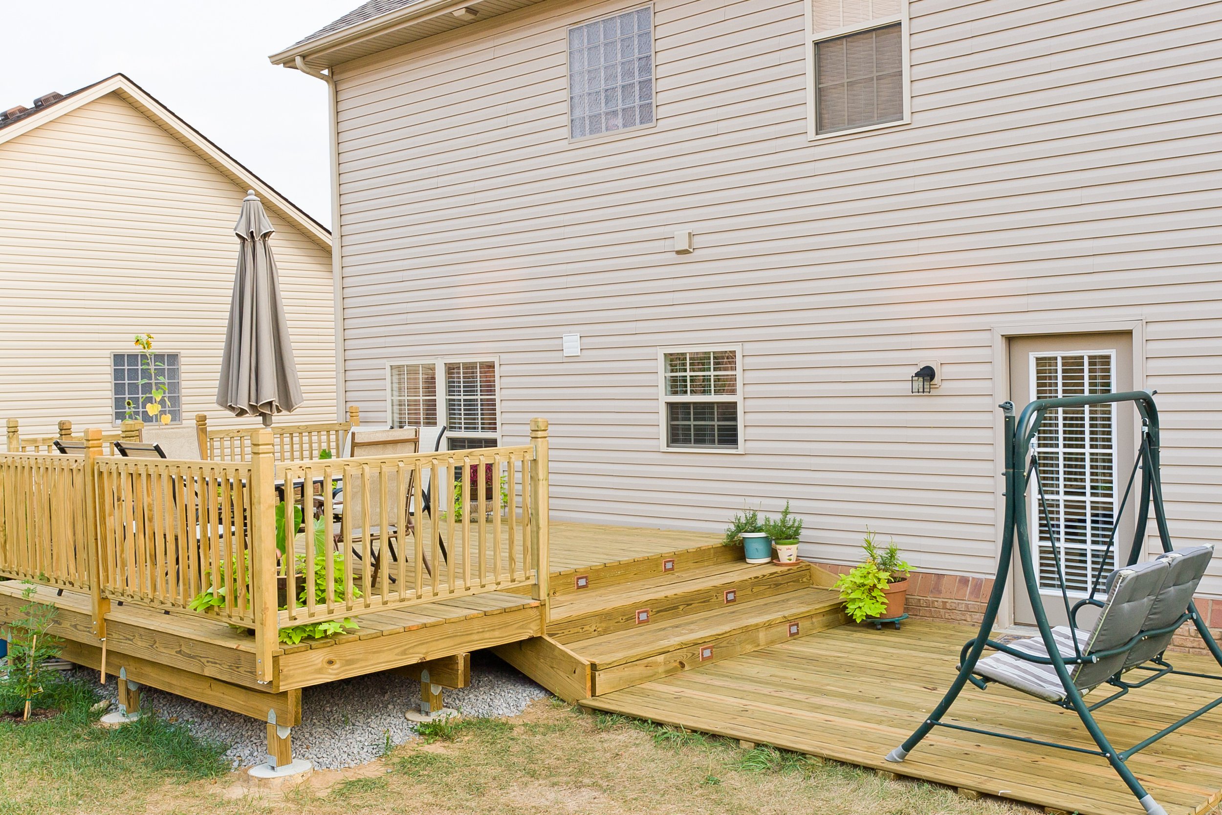 Backyard with a wooden patio, outdoor furniture, patio swing, potted plants, and a black lantern wall light attached to the house.