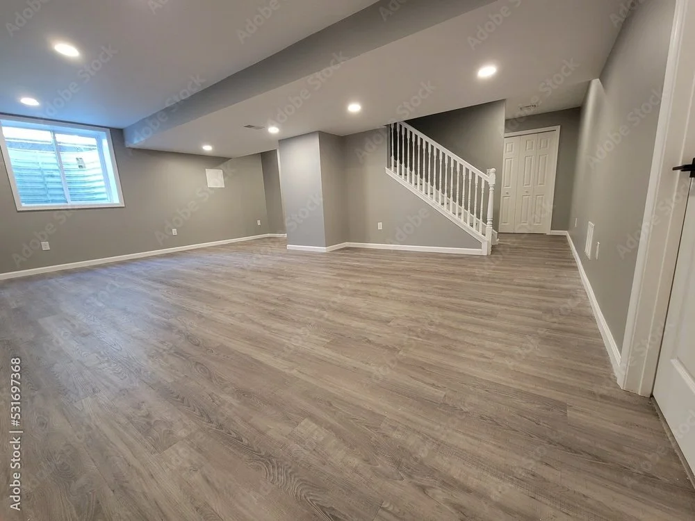Empty basement room with gray walls, wood flooring, white baseboards, a window, and a staircase with white railing.