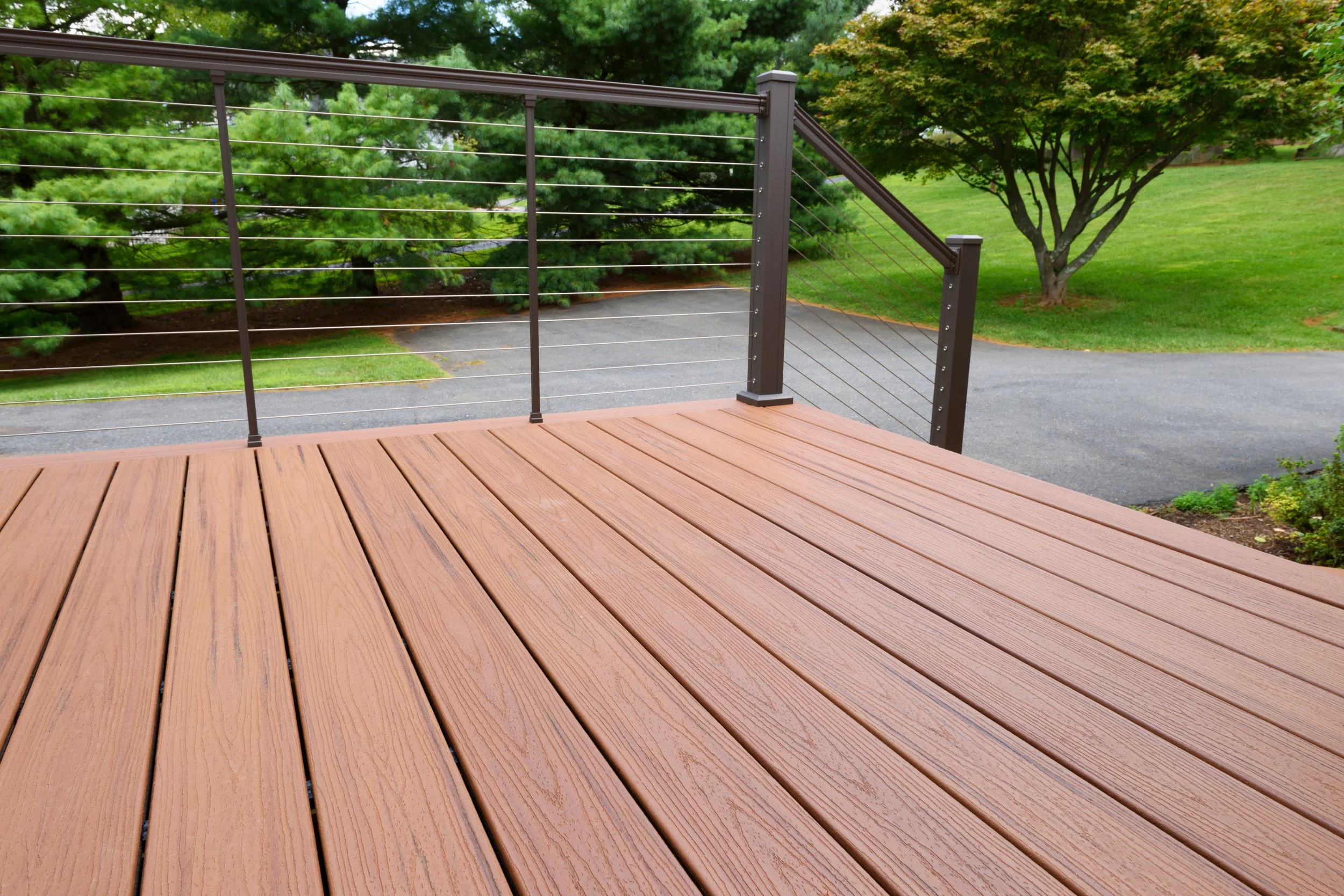 A wooden deck with metal railing overlooking a driveway, with green trees and grass in the background.
