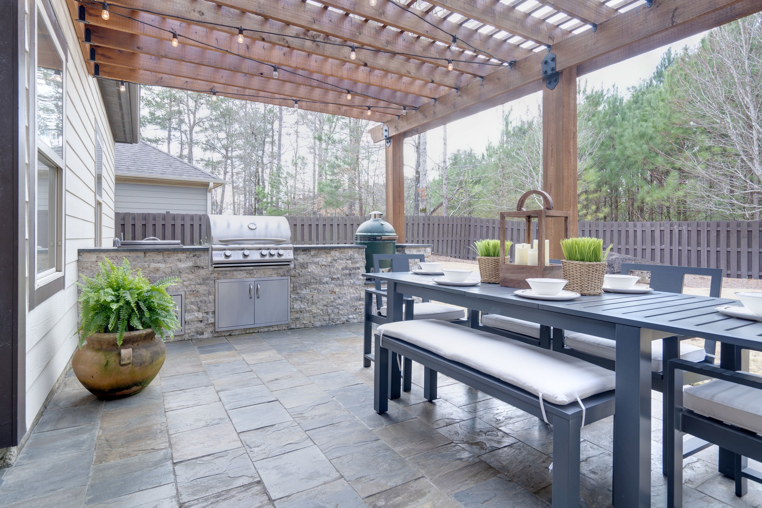 Outdoor patio with a dining table and benches, a grill area with a built-in grill, a potted fern, decorative lanterns, and string lights under a wooden pergola, surrounded by a wooden fence and trees.