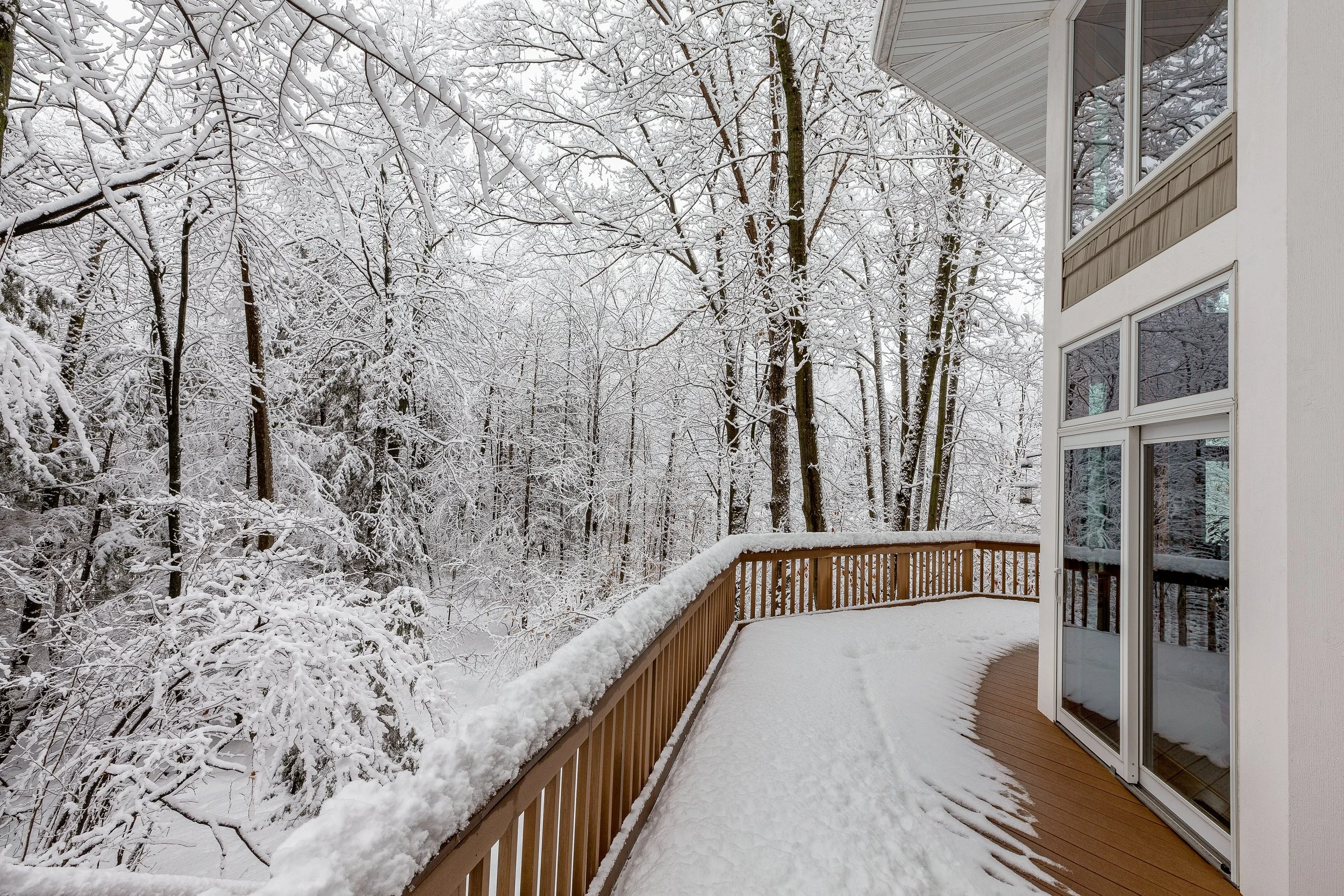 Snow-covered outdoor balcony with railing and wooded forest in the background, seen through large windows of a house.