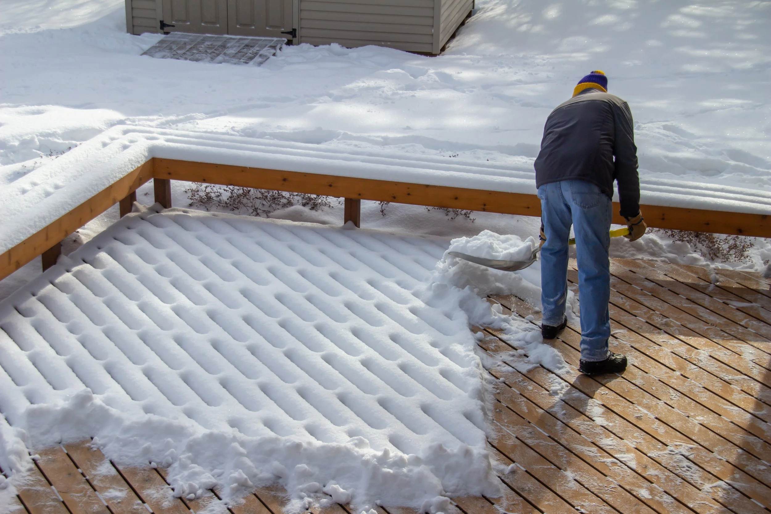 A person shoveling snow off a wooden deck in a residential backyard during winter.