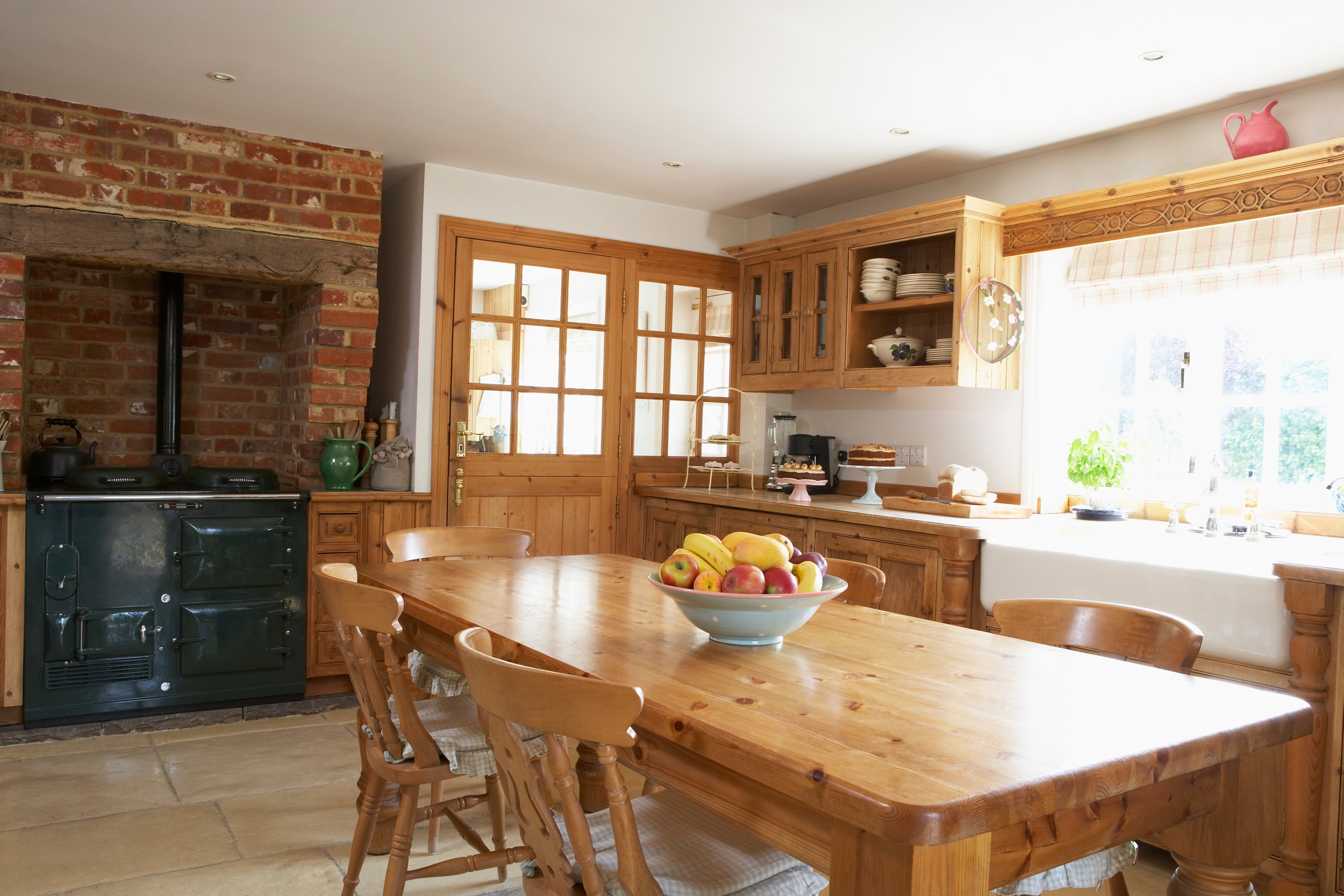 Kitchen with wooden cabinets, brick fireplace, wooden table with chairs, window with checkered curtain, and a bowl of apples and bananas on the table.