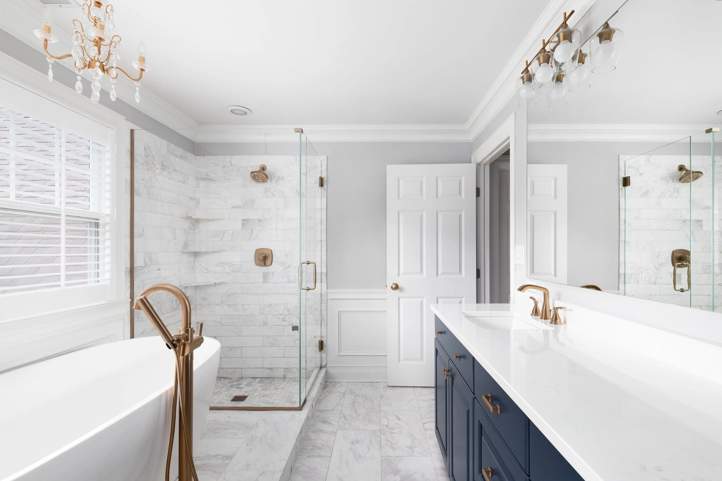 Bathroom with white marble floors and walls, a glass-enclosed shower, a white bathtub with bronze fixtures, a dark blue vanity with bronze handles, a large mirror, and a window with white blinds.