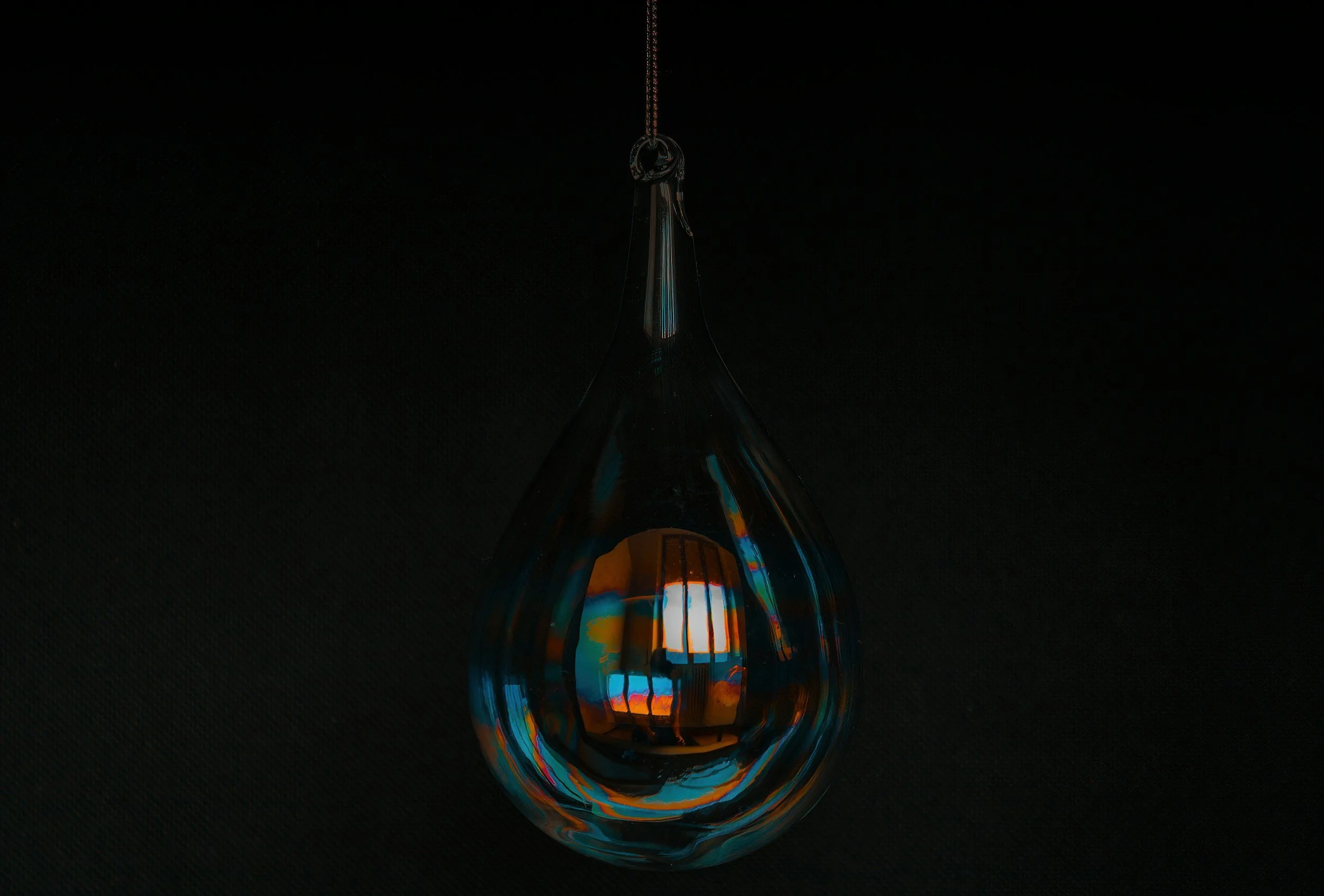 A glass teardrop-shaped ornament hanging by a string, reflecting a colorful window and interior lights against a black background.