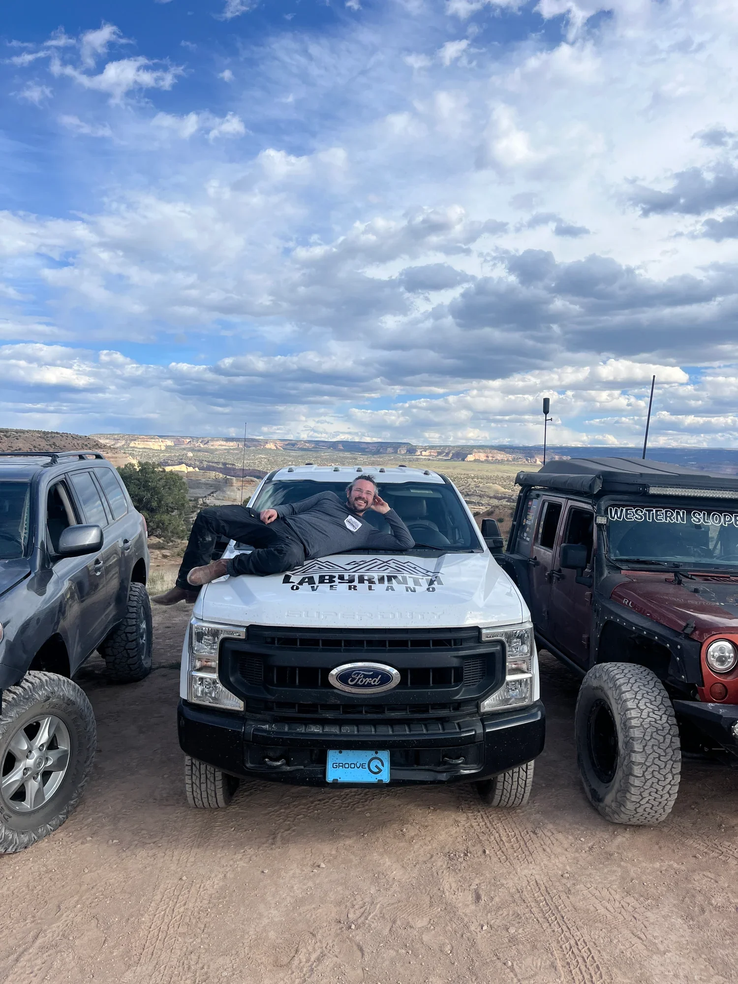 Man lying on the hood of a white Ford vehicle, smiling, with a dirt landscape and cloudy sky in the background, flanked by off-road vehicles.